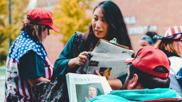 Kvinne holder en avis på gaten fremfor en mann som sitter og leser en avis