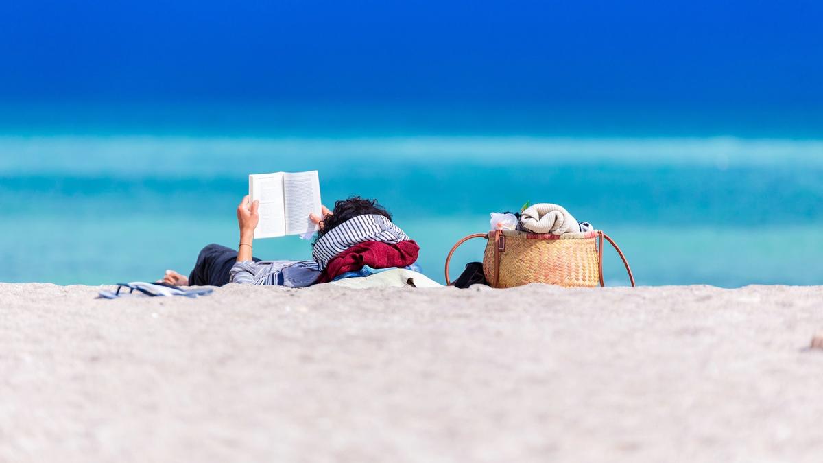 Illustrasjonsbilde av en person som ligger å leser en bok på en badestrand med blågrønt hav i bakgrunnen. Bildet tatt av Dan Dumitriu fra Unsplash