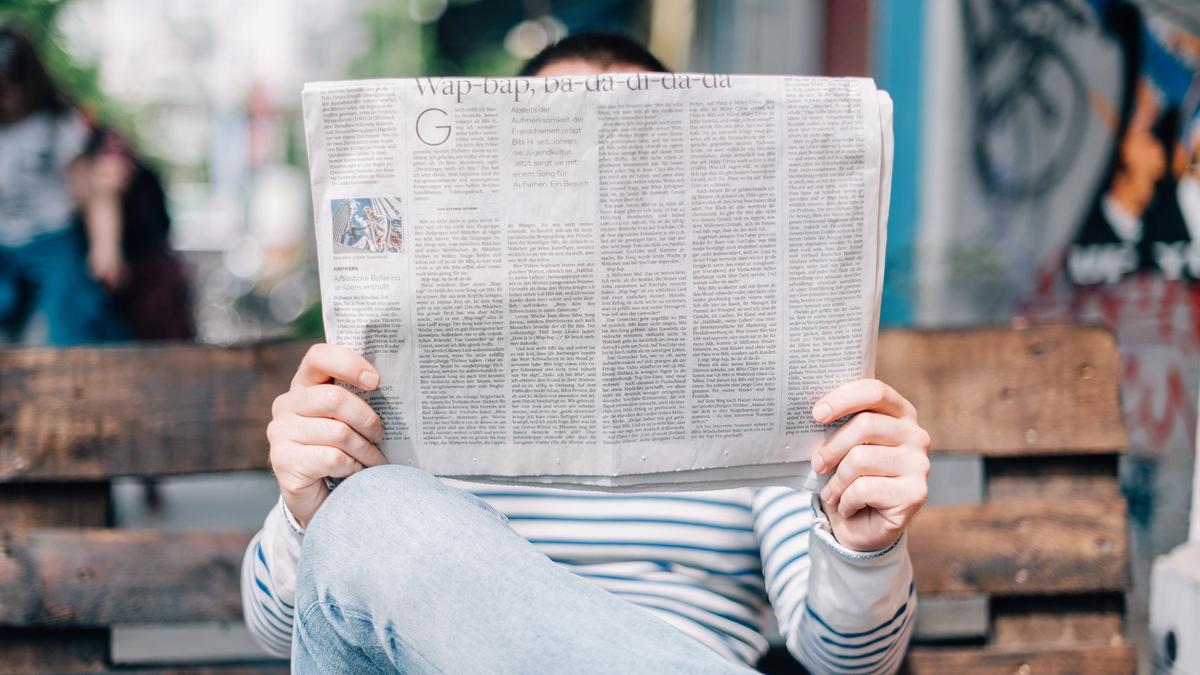 Mann med lys dongribukser og hvit genser med blå striper sitter på en brun benk og leser avisen. Illustrasjonsbilde tatt av Roman Kraft fra Unsplash