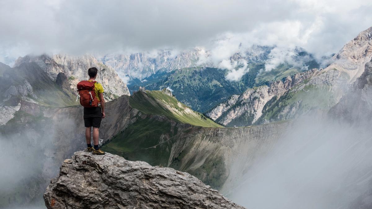 man with red backpack standing on cliff facing mountains under white fog