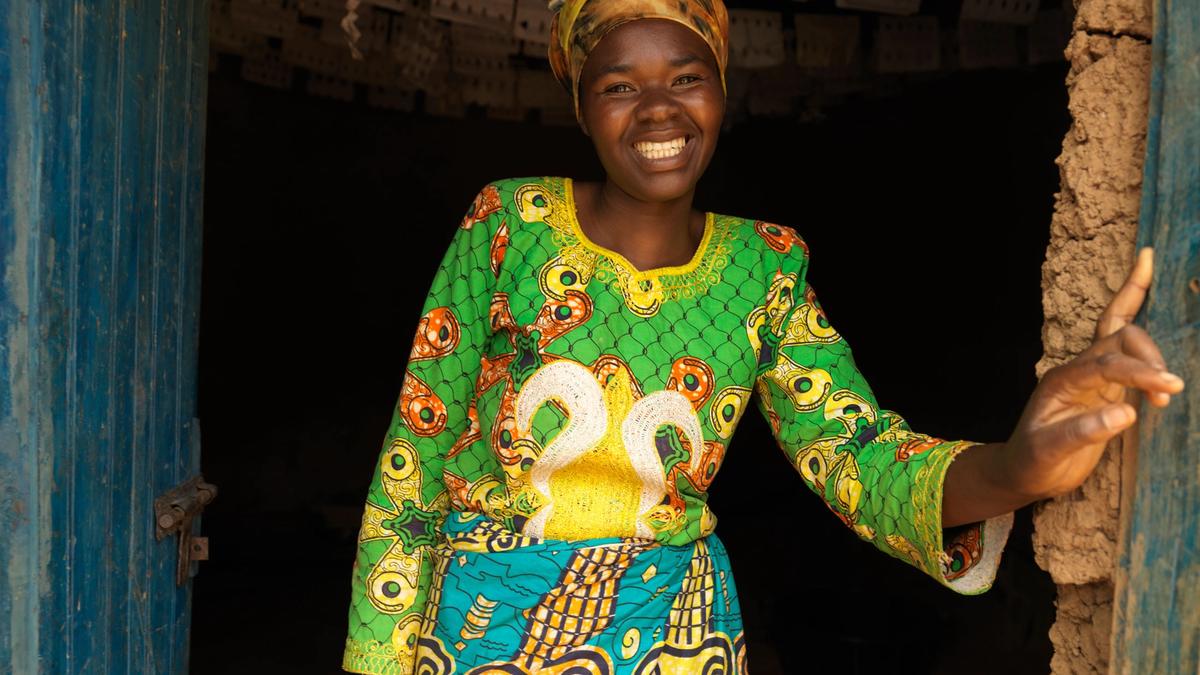 En kvinne som står i en døråpning og smiler. Hun har på seg grønn fargerik bluse og et turkis skjørt. Bilde tatt i Rawanda av Care. Publisert med tillatelse fra Care.