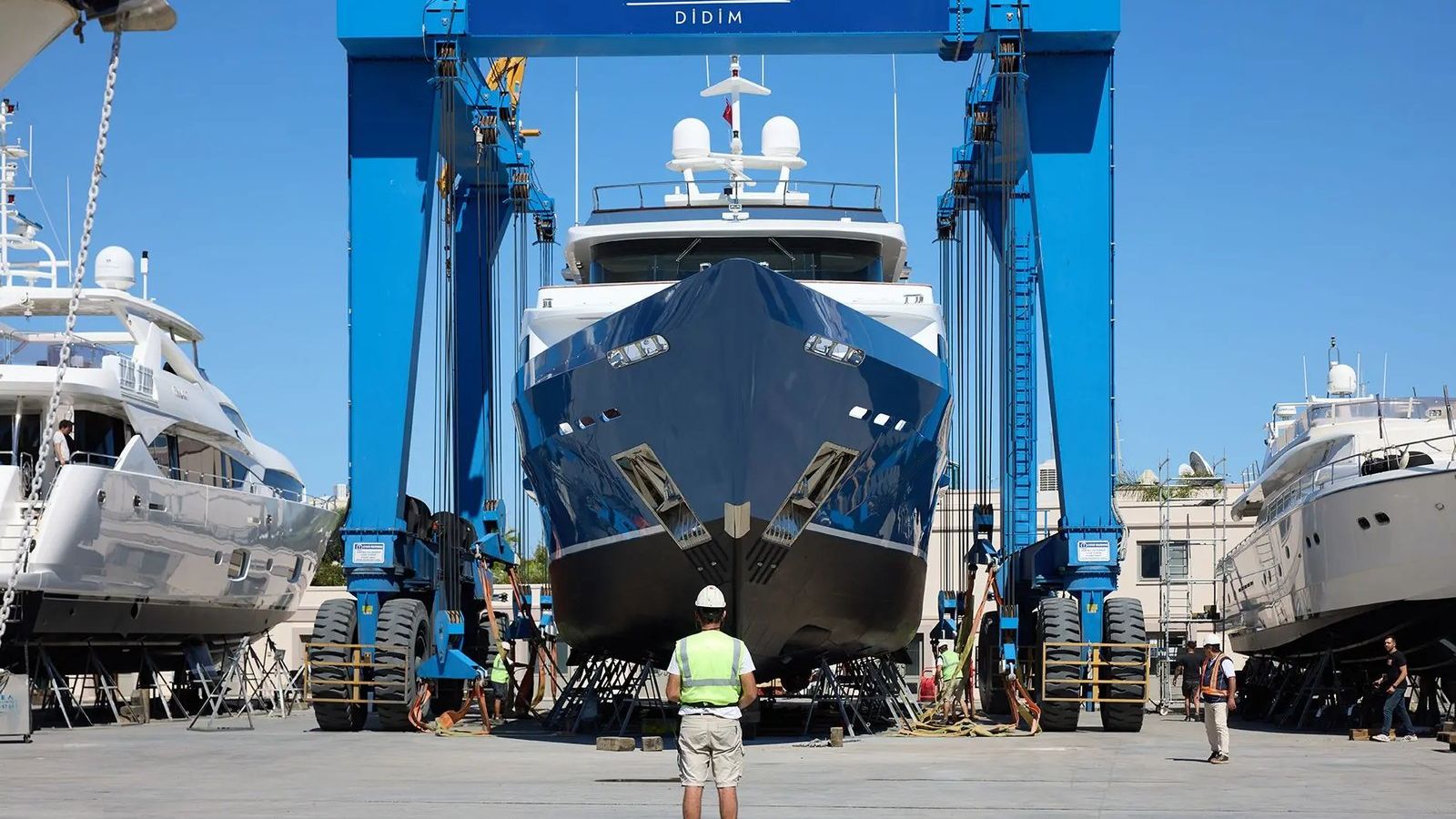 A marina worker in a high-visibility vest standing in front of a superyacht suspended in a blue travel lift at D-Marin Didim