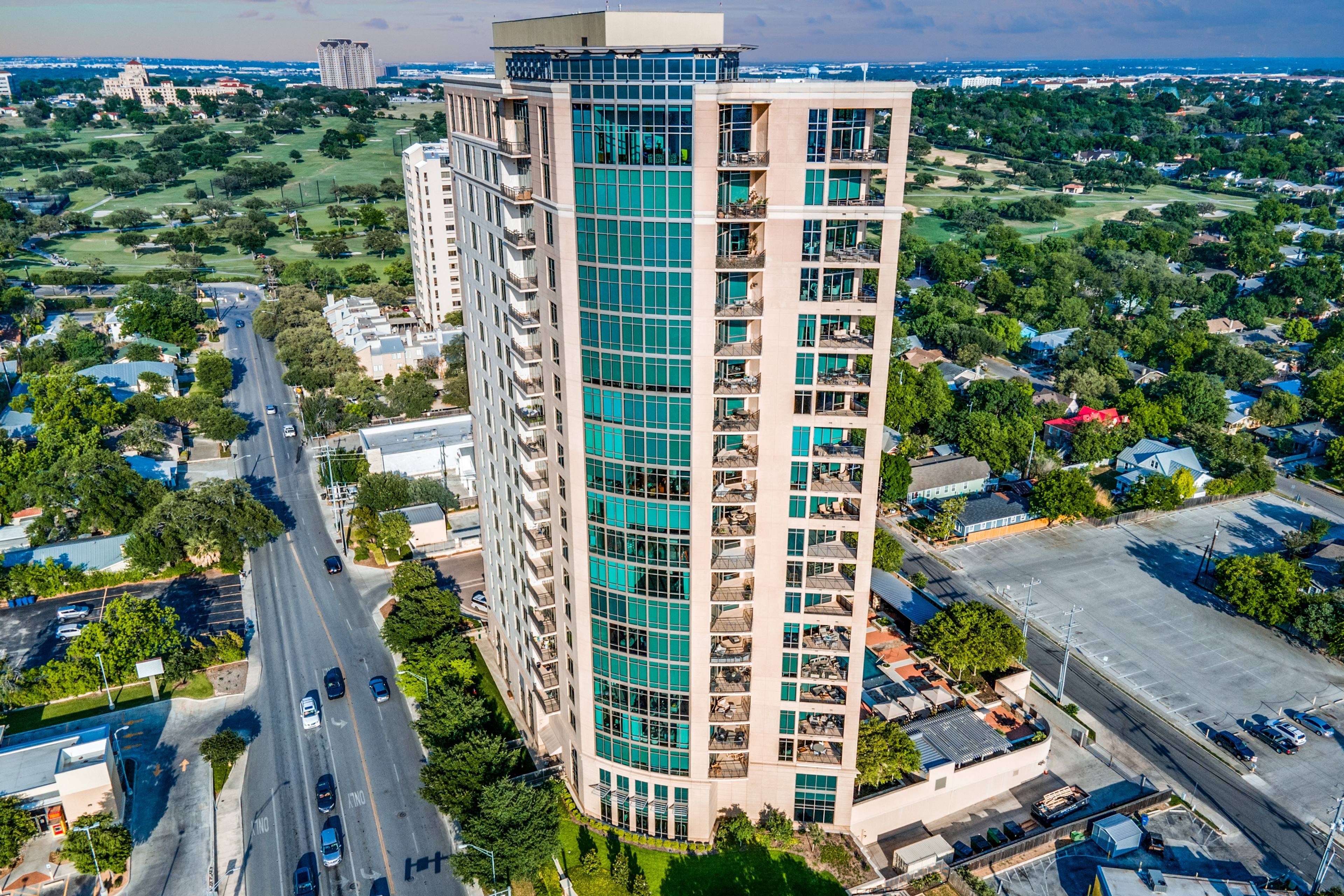 Aerial drone photograph of a luxury glass high-rise apartment tower with golf course views and tree-lined neighborhood in San Antonio, Texas