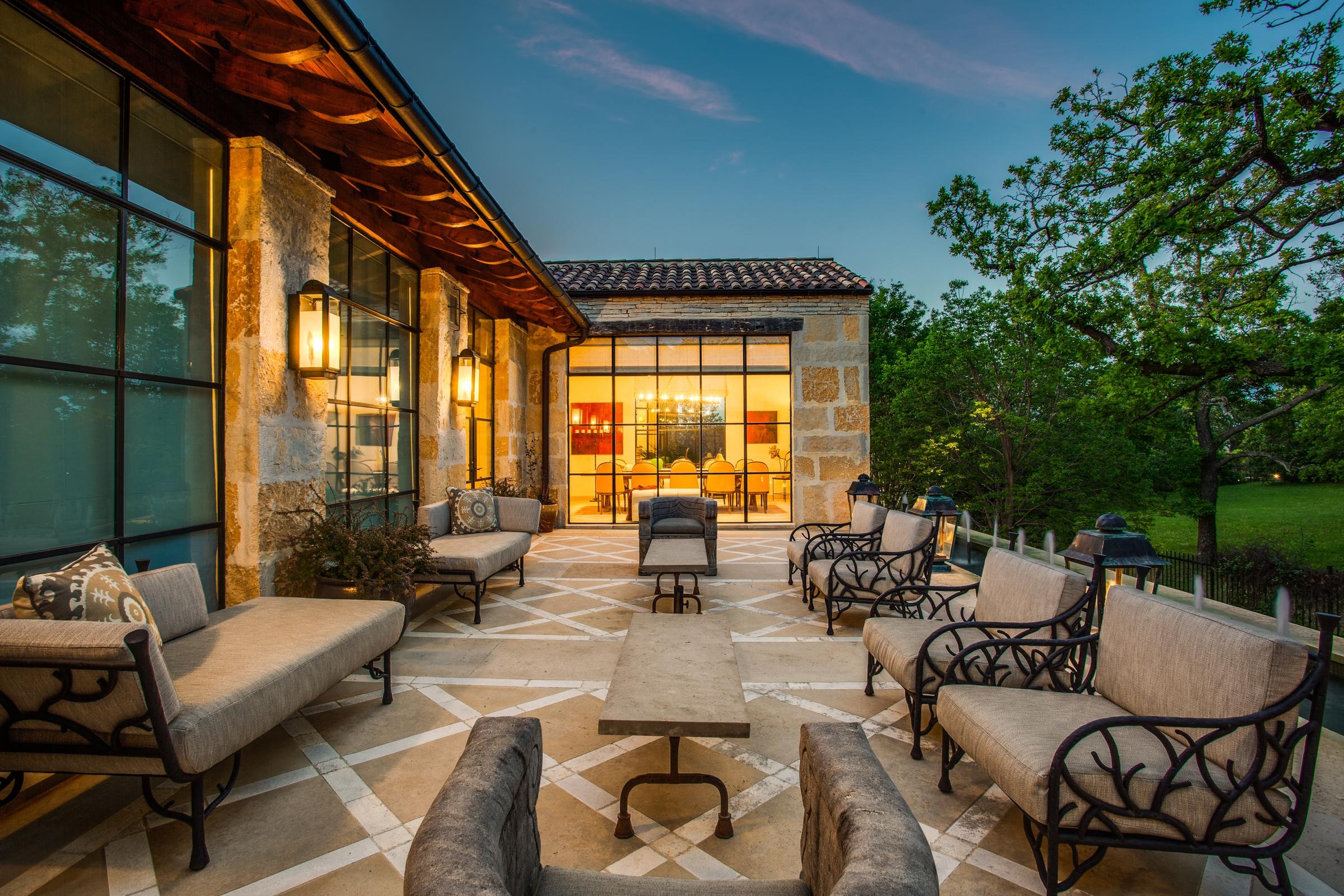 Twilight view of a stone patio with wrought iron seating and warm interior lighting at a luxury Westlake, Texas home