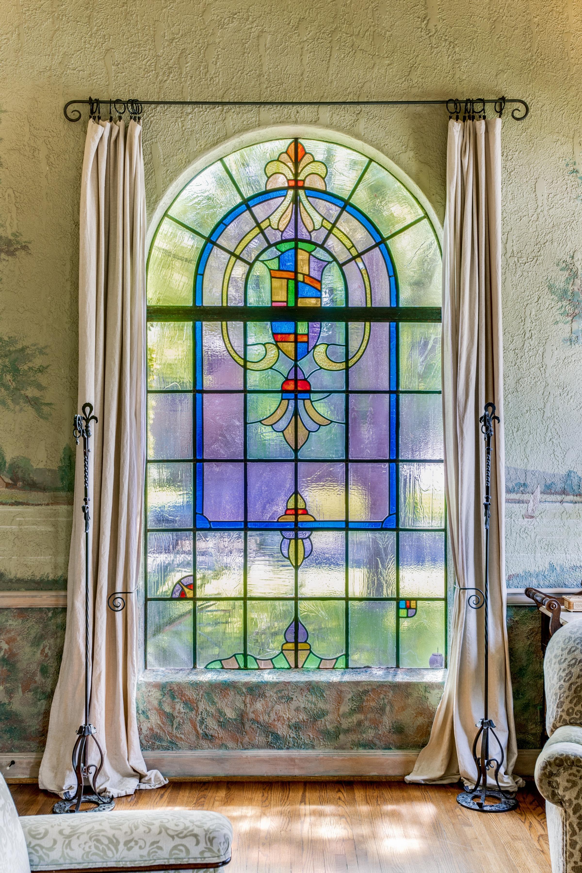 Arched leaded stained glass window with curtains, plaster walls, and painted landscape mural trim in a historic Texas estate.