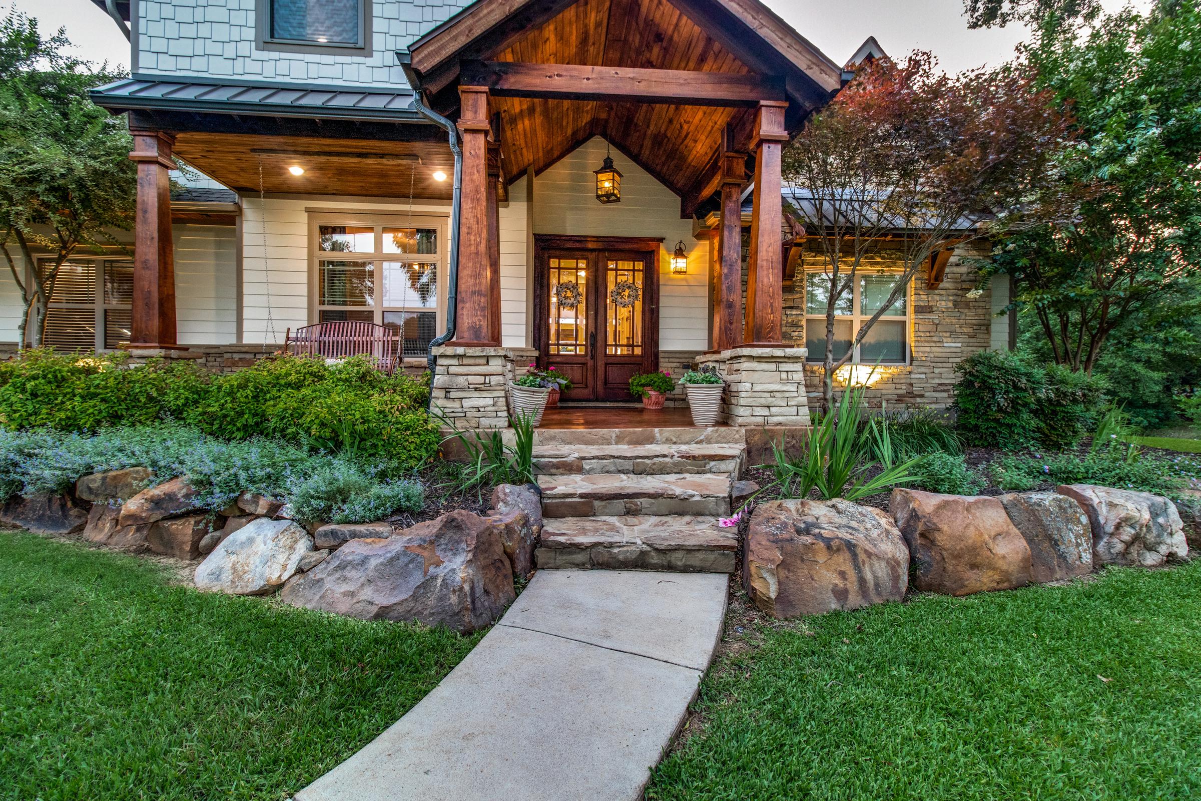 Craftsman-style covered porch entry with timber beams, stone columns, and landscaped steps lit at dusk in Bells, Texas