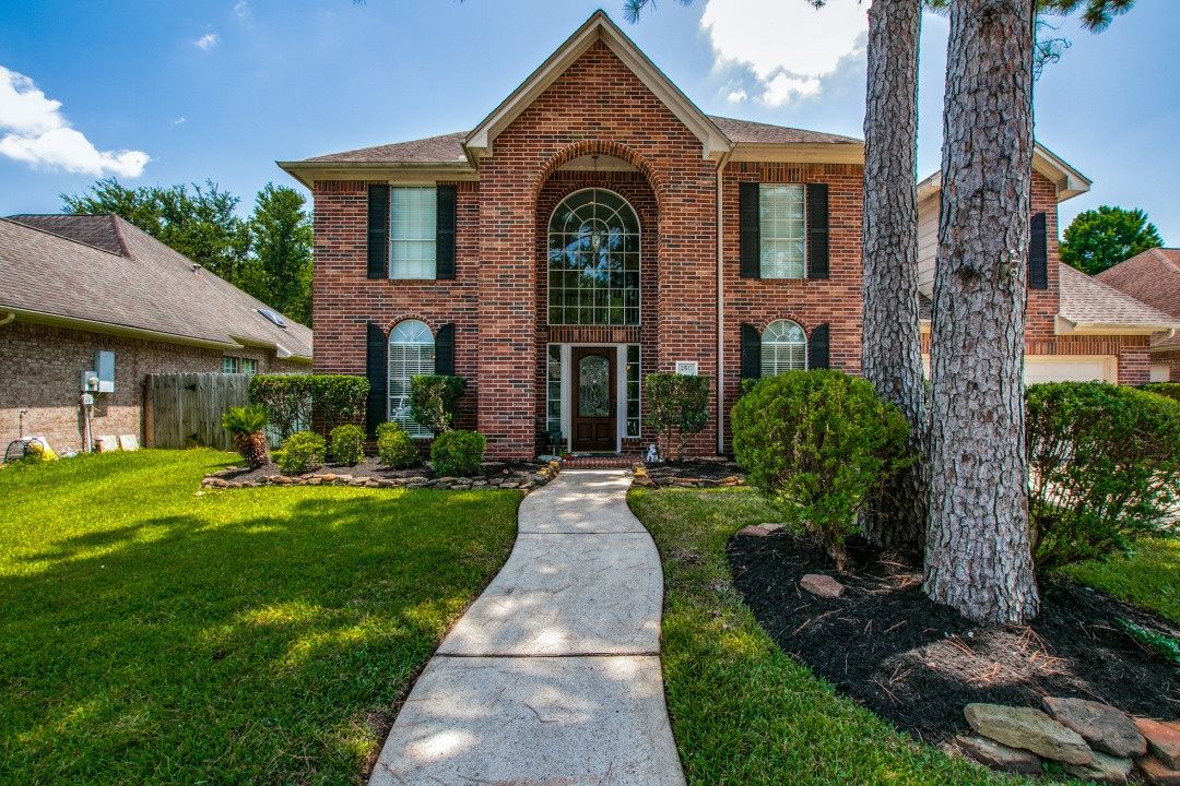 Two-story brick home with sidewalk approach, large pine trees, and manicured green lawn in Pearland, Texas