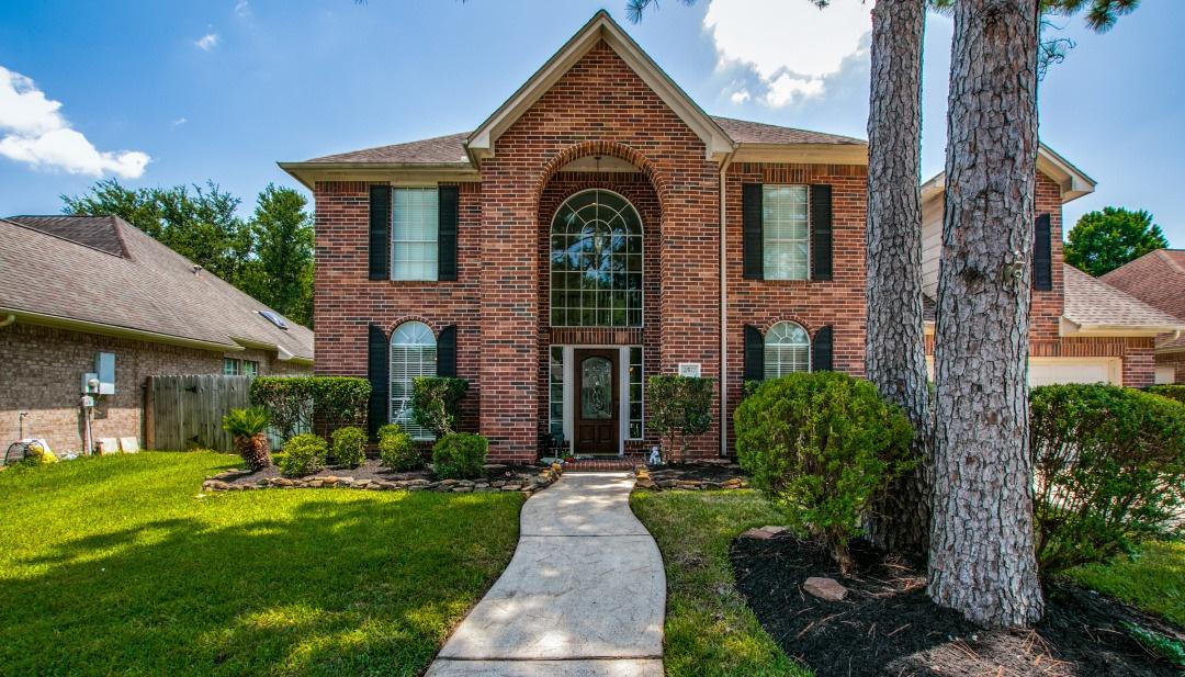 Two-story brick home with sidewalk approach, large pine trees, and manicured green lawn in Pearland, Texas