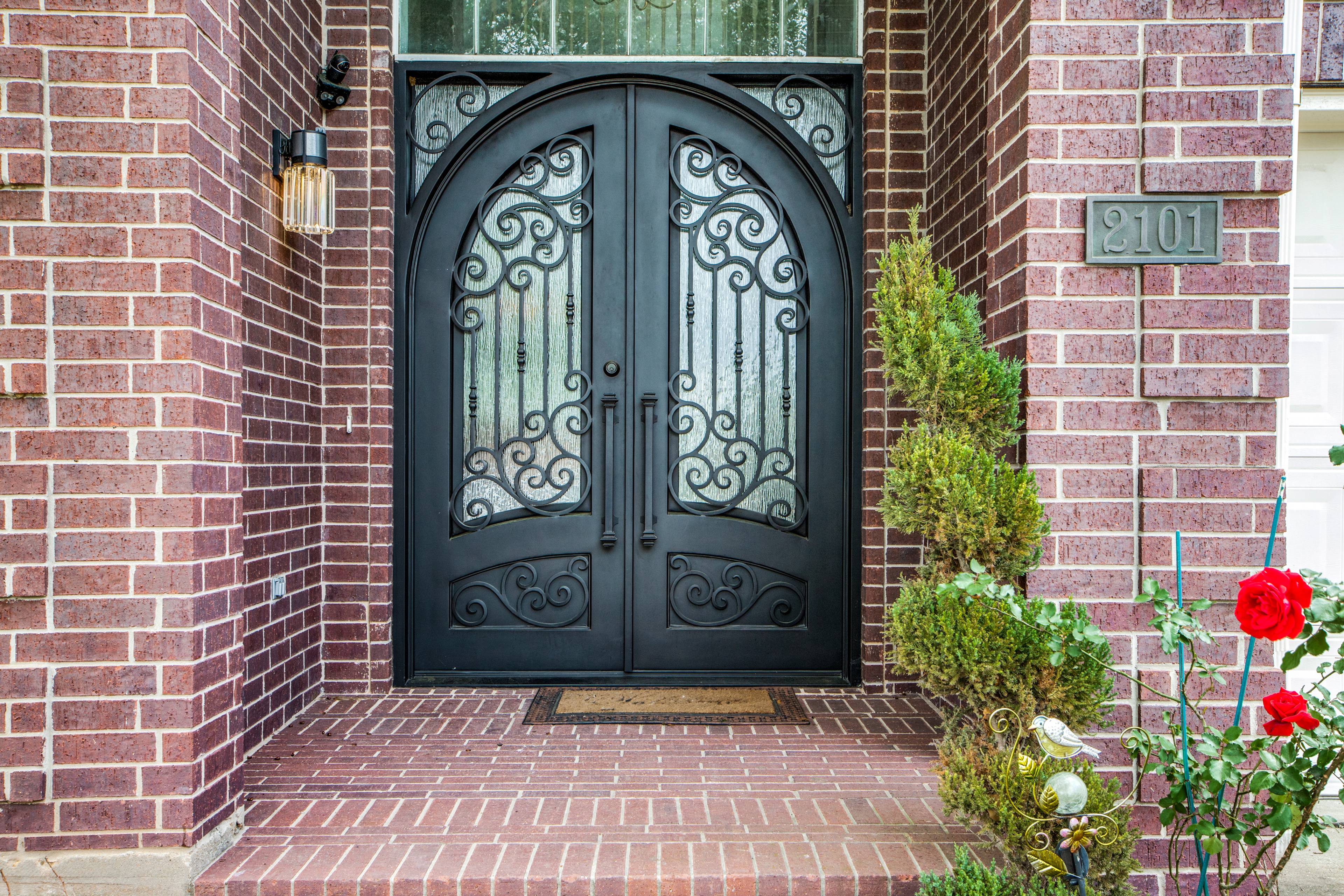 Front entry detail of a Pearland, TX brick home with arched black wrought-iron double doors, scrollwork panels, seeded glass, a vintage lantern sconce, and a potted rose bush on the porch.