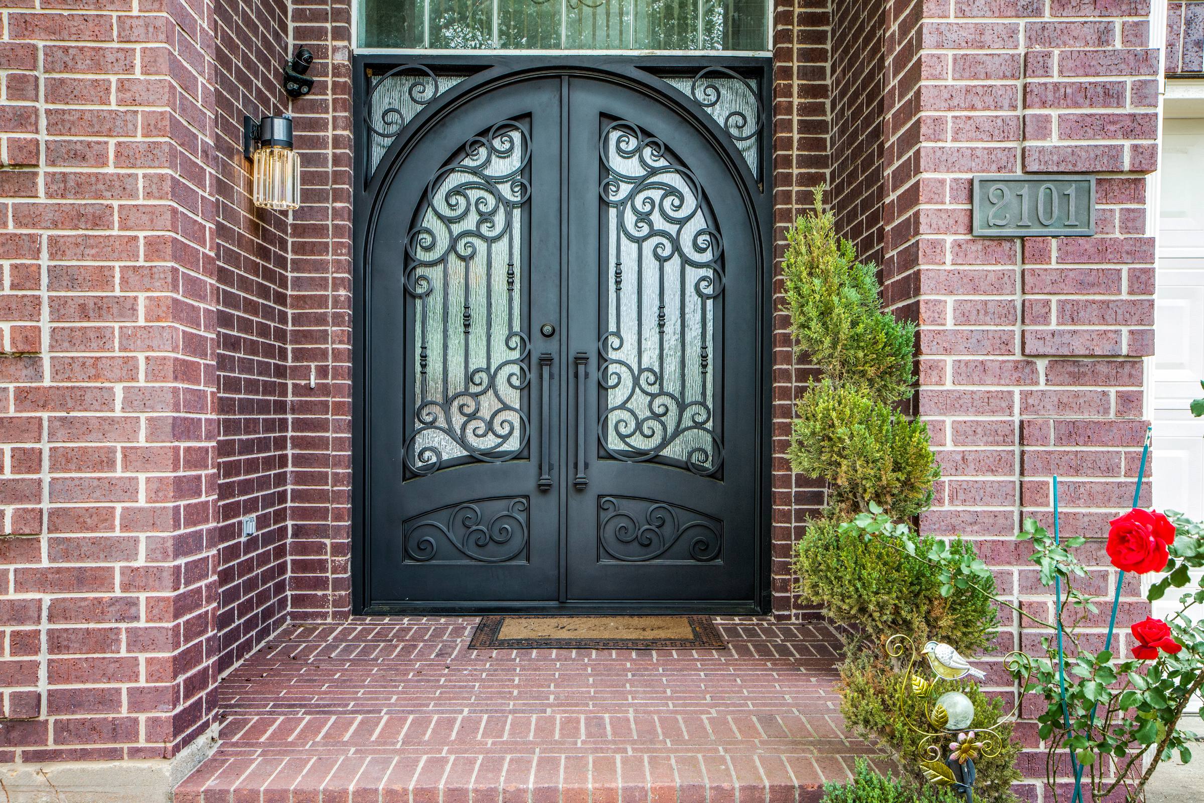 Front entry detail of a Pearland, TX brick home with arched black wrought-iron double doors, scrollwork panels, seeded glass, a vintage lantern sconce, and a potted rose bush on the porch.