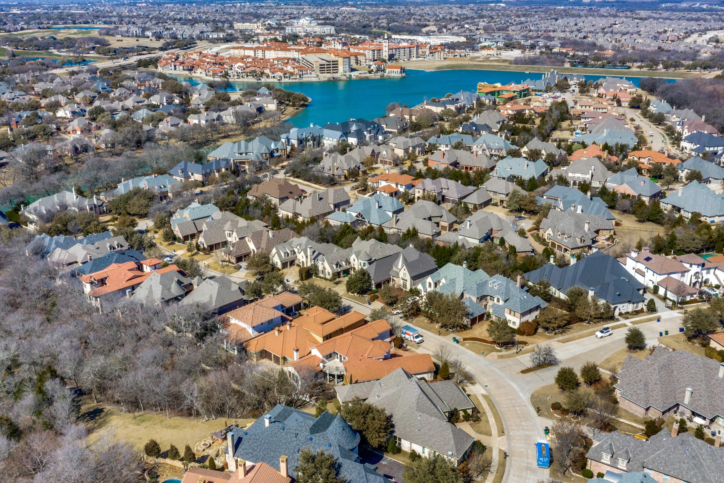Aerial drone view of an established lakefront residential community with tile-roof luxury homes and a turquoise lake in DeSoto, Texas