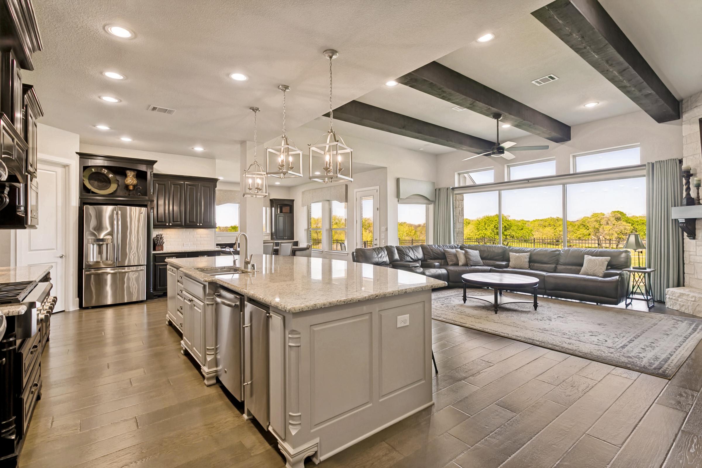 Open-concept kitchen and living room with a granite island, wood beams, and wall of windows to green space in a Prosper home