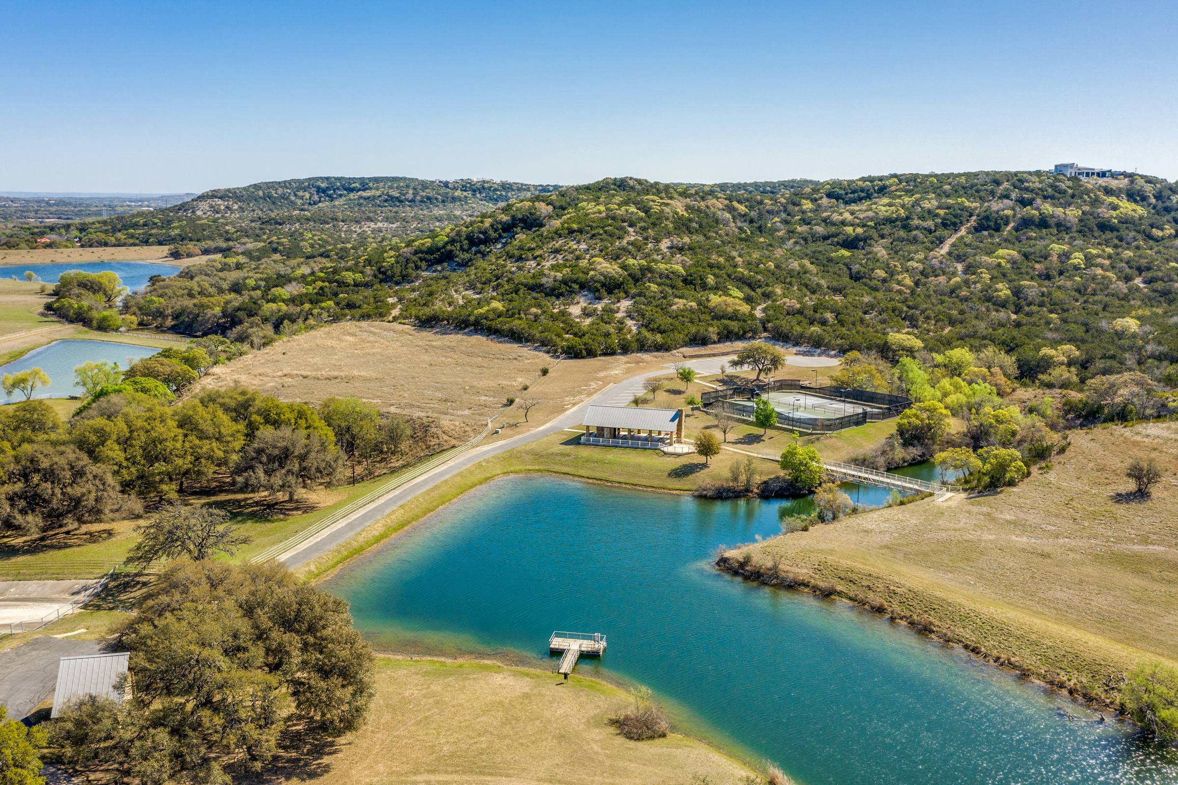 Drone aerial of a Texas Hill Country community with a stocked pond, fishing dock, sport courts, and stone pavilion among wooded hills in Boerne
