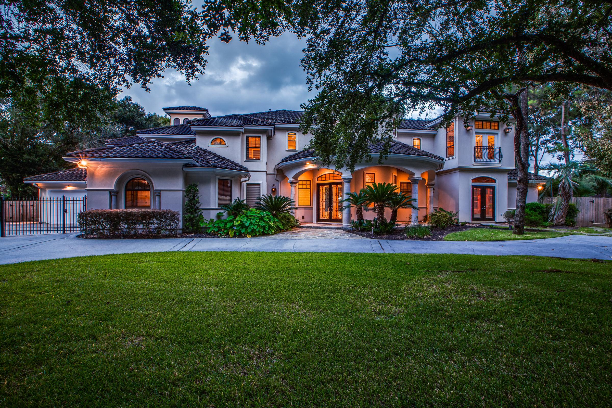Mediterranean-style stucco estate with a tile roof lit at dusk beneath mature oak trees in Spring, Texas