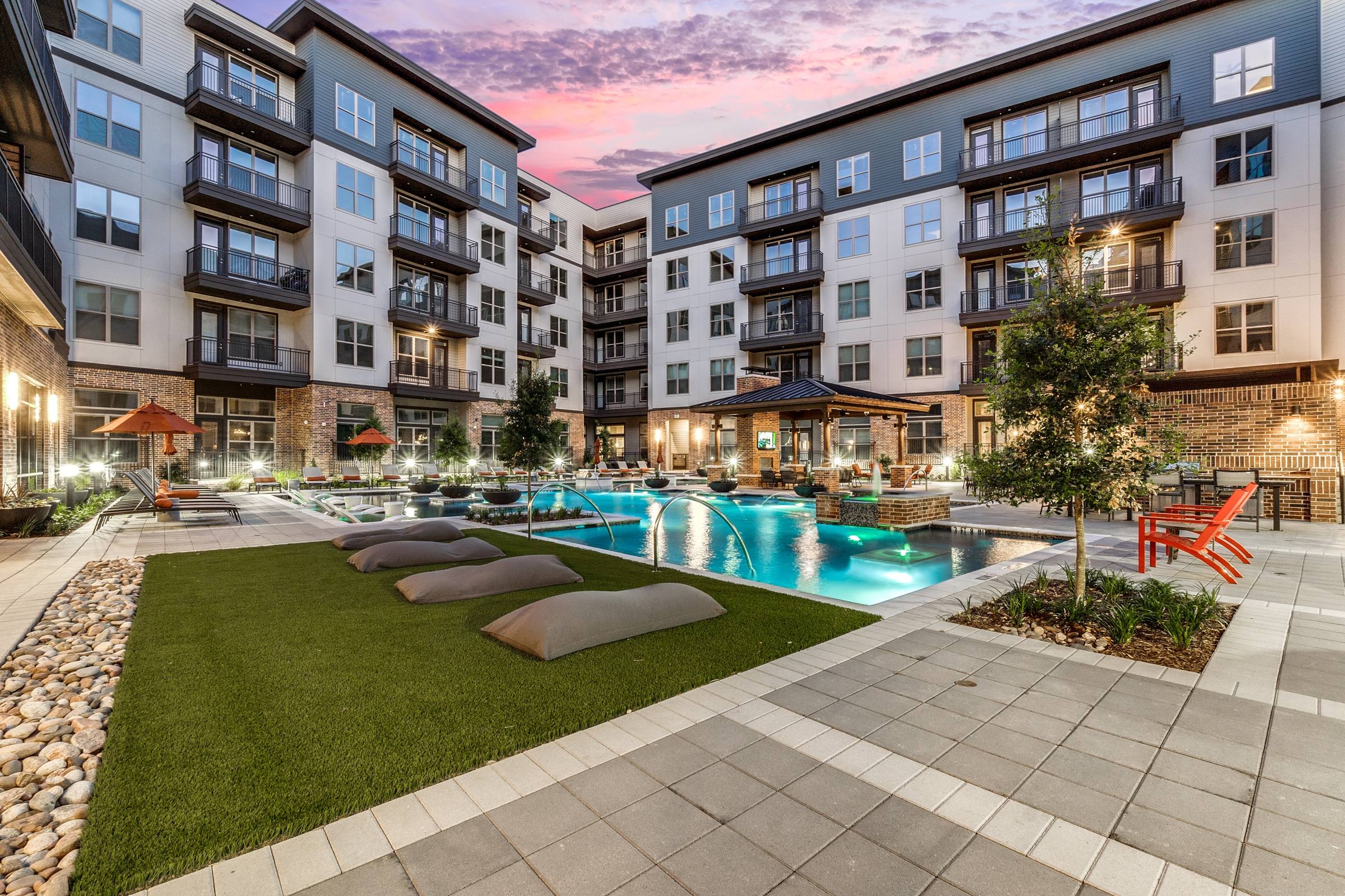 Illuminated pool with fountains and turf lounge area at sunset at a modern apartment community in Dallas, Texas