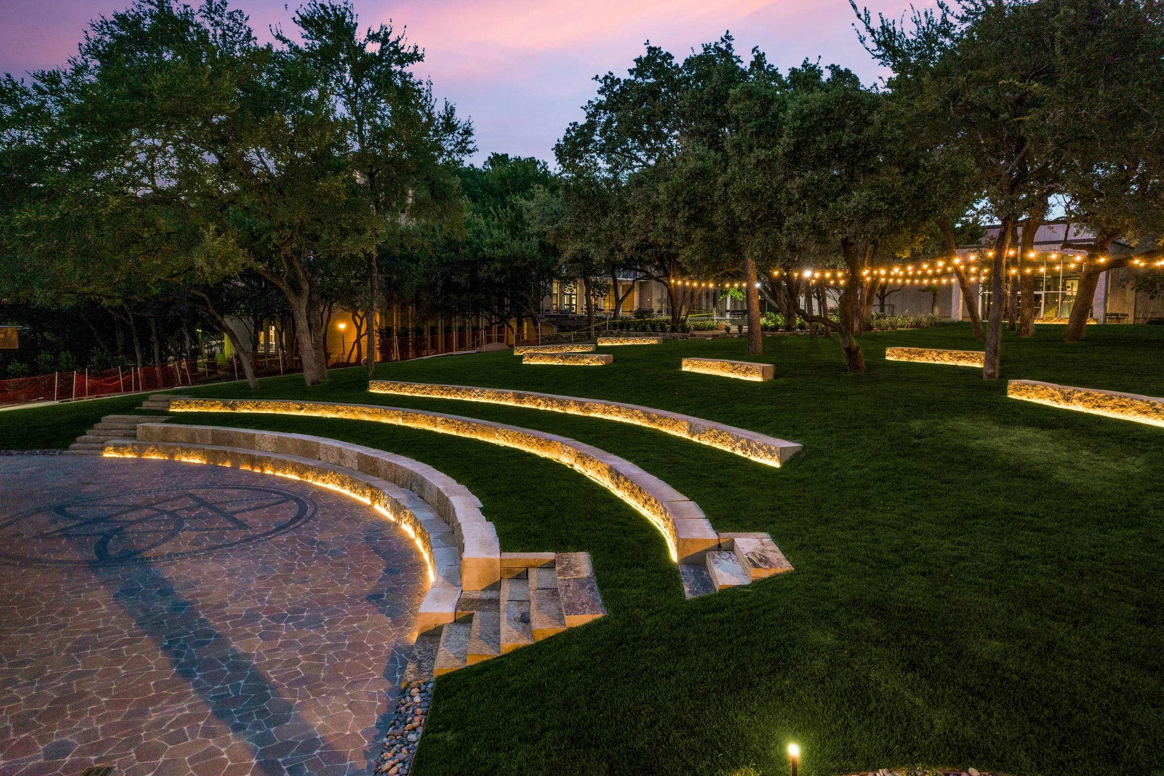 Landscaped outdoor amphitheater with illuminated stone terracing and string lights at twilight in Irving, Texas