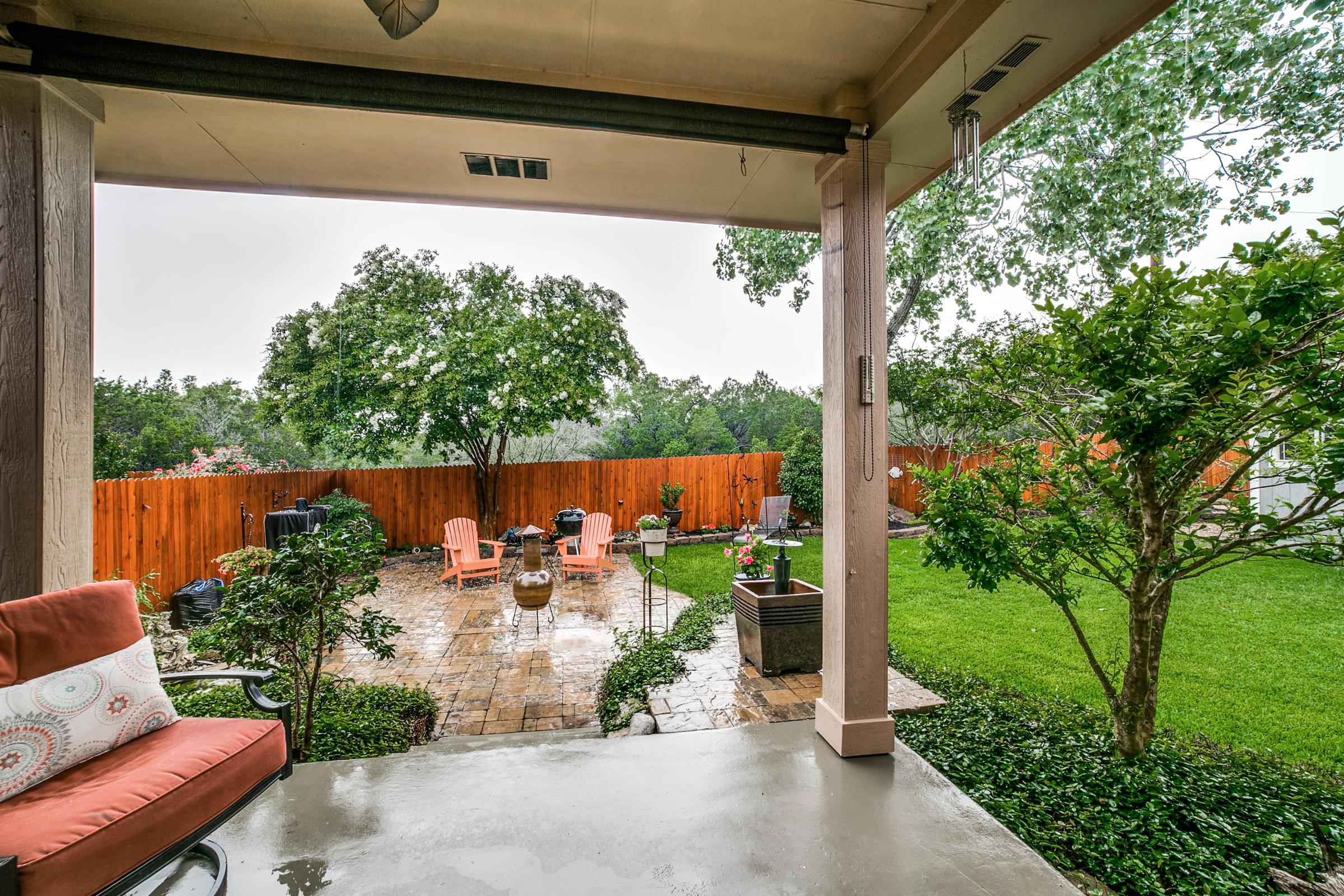Covered patio looking onto a lush backyard with paver stone area, orange Adirondack chairs, and wooden privacy fence on a rainy day in San Antonio, TX