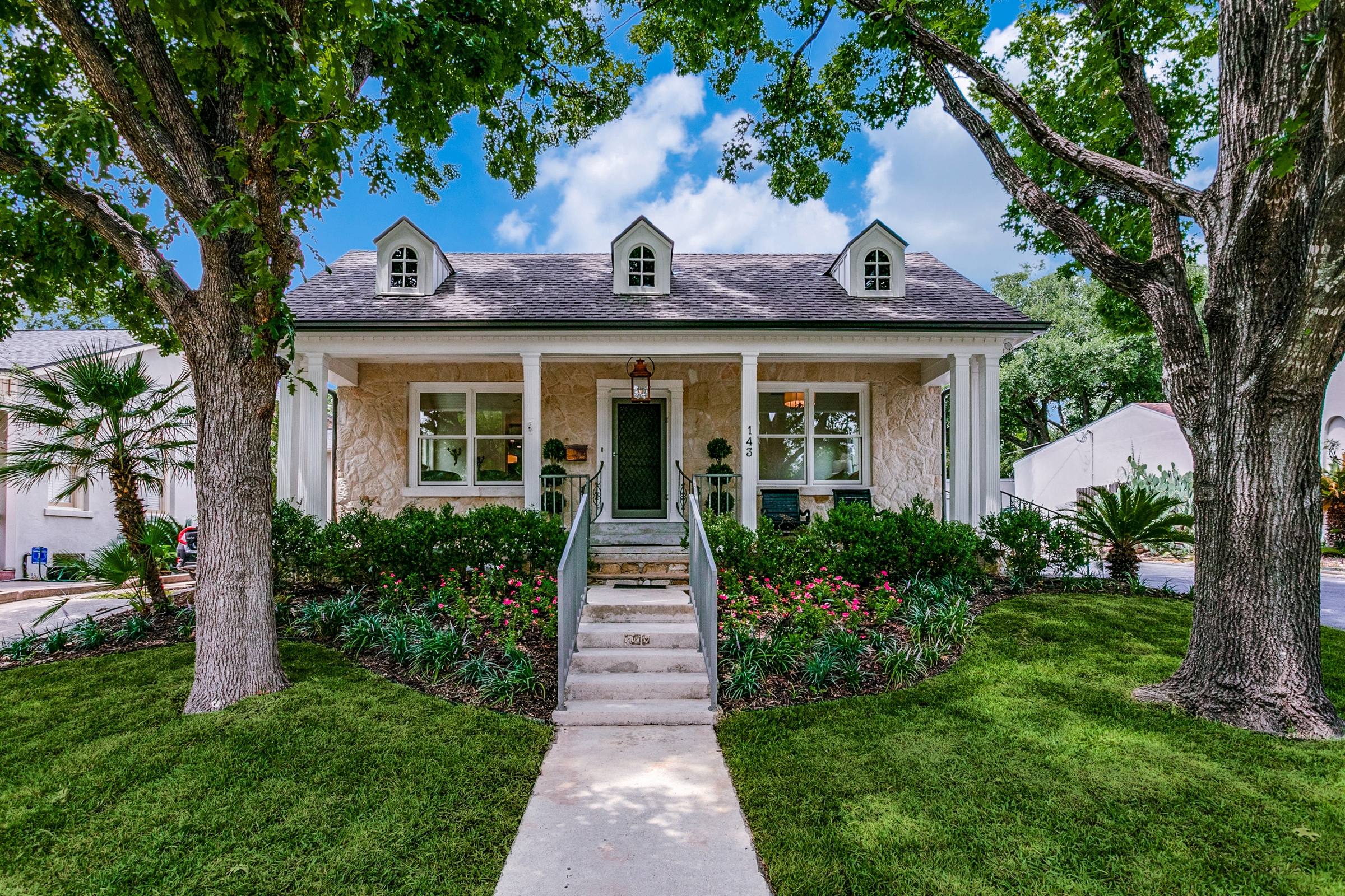 Charming cottage-style home with covered front porch, dormer windows, and lush landscaping in Alamo Heights, Texas