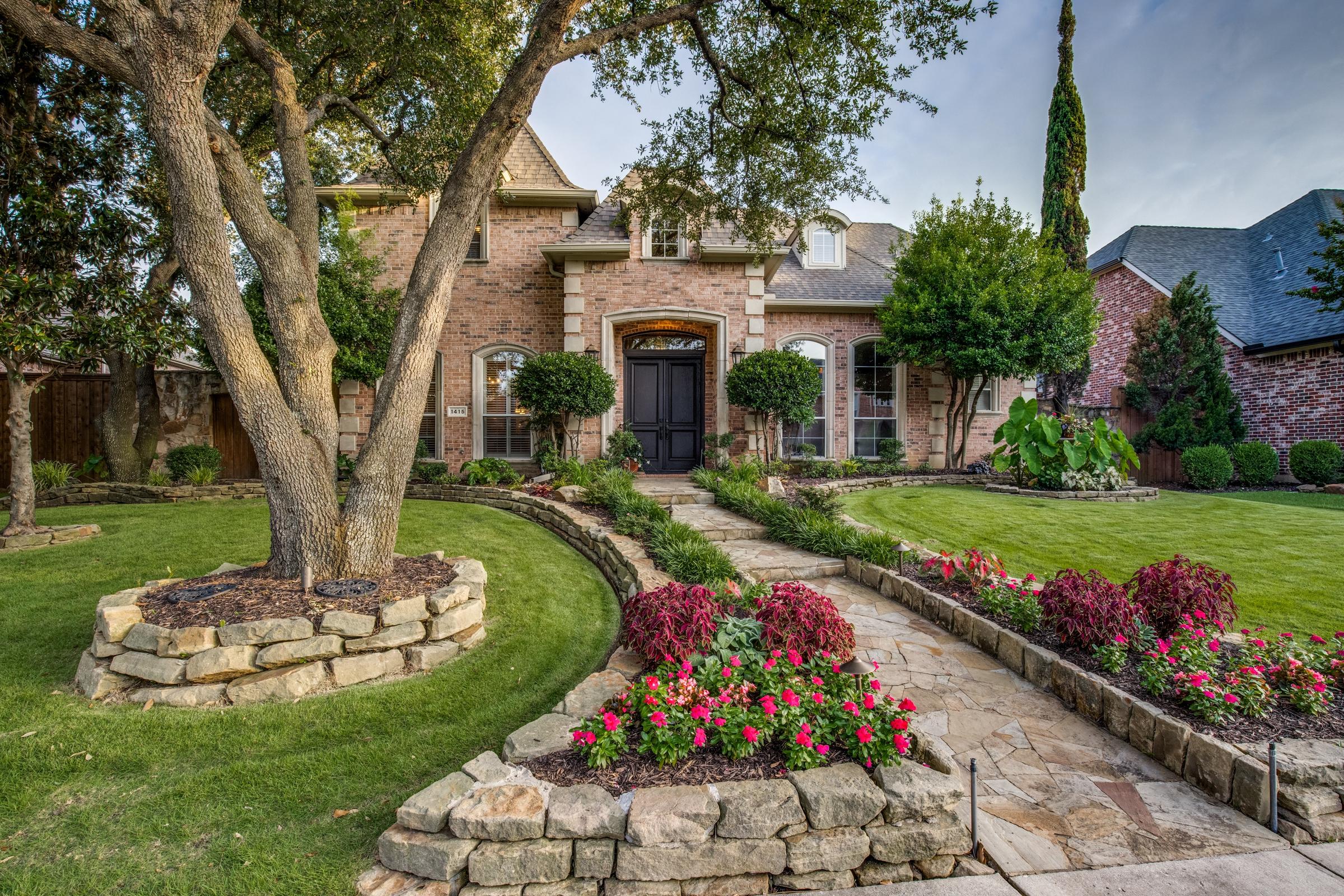 Brick home with colorful spring flower beds, stone pathway, and mature oak trees at dusk in Coppell, Texas
