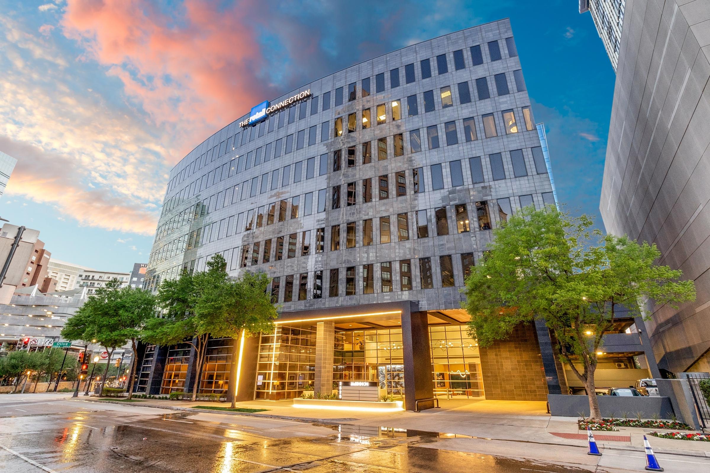 Modern curved glass office building with illuminated lobby and wet pavement reflecting twilight colors in Dallas, Texas
