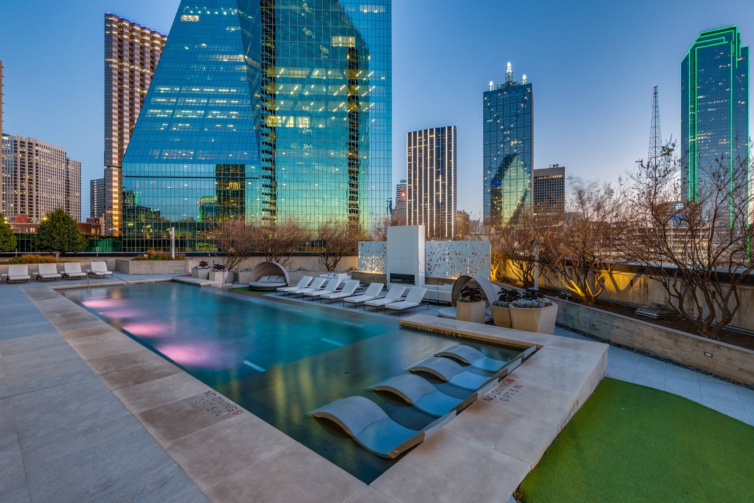 Luxury apartment rooftop pool deck with in-water loungers and downtown Dallas glass skyline reflected at twilight