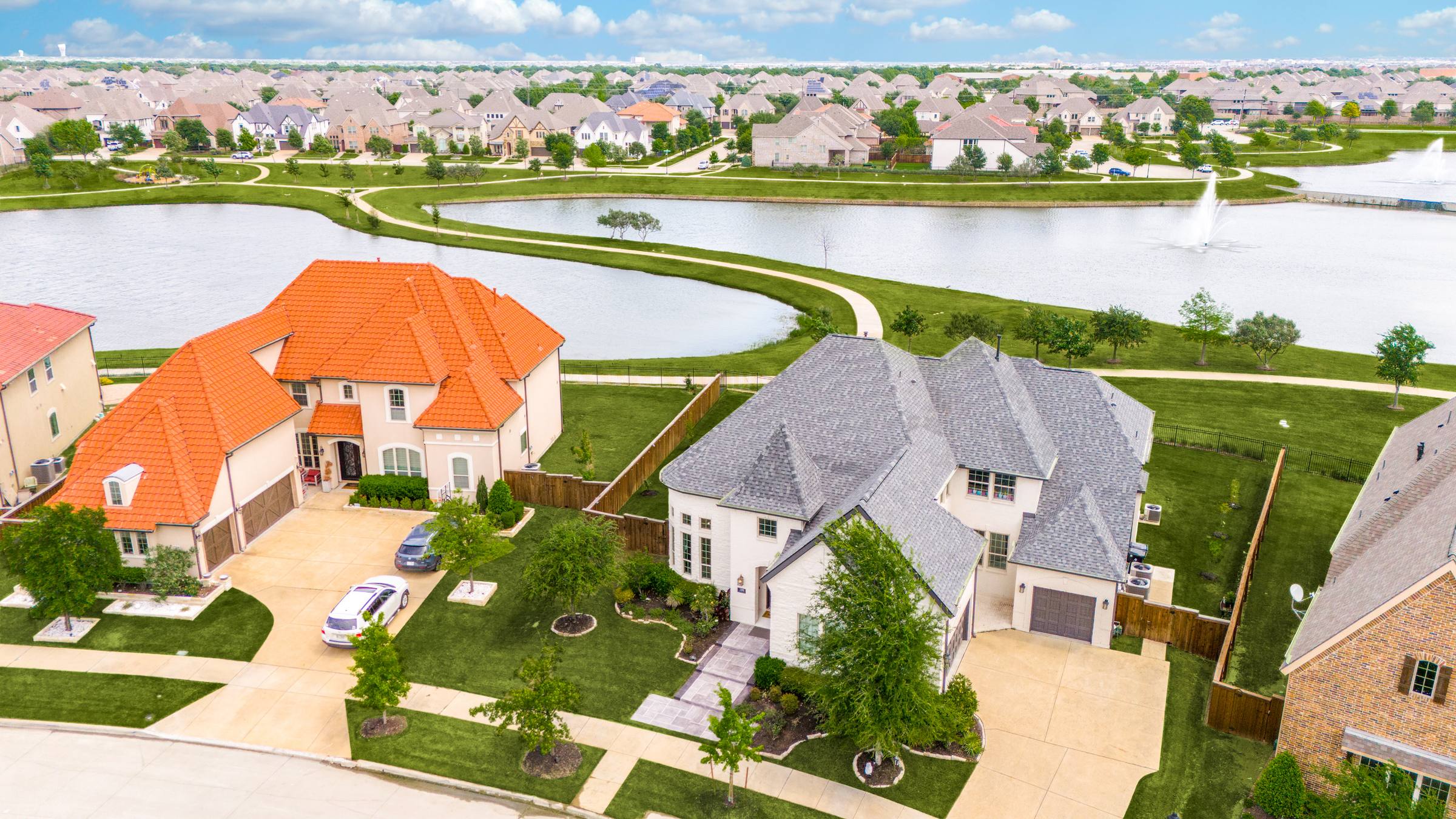 Aerial of a Frisco, TX lakeside suburban street with two custom homes, a community walking-path lake, and rooftops to the horizon — enhanced with Shoot2Sell's Aerial Green Grass treatment for vibrant lawn color.