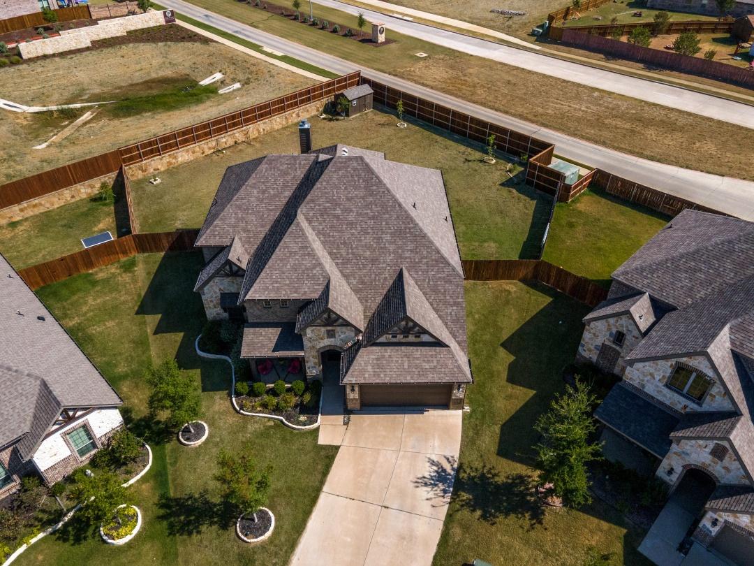 Aerial drone photograph of a new construction stone and brick single-family home with three-car garage and fenced backyard in a suburban Burleson, Texas neighborhood