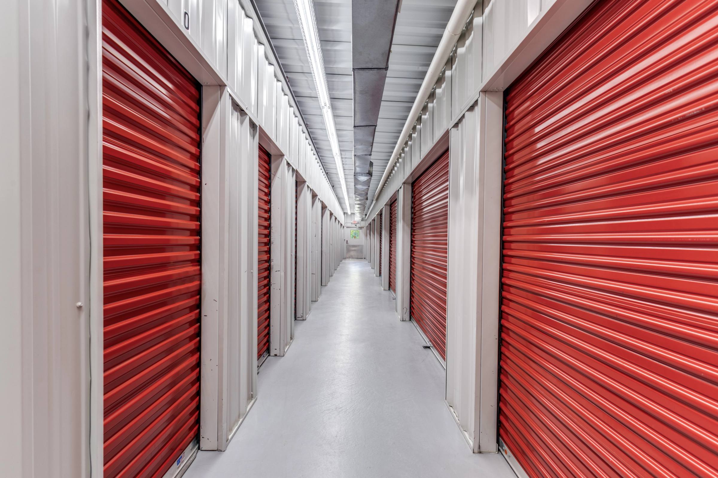 Interior corridor of a climate-controlled self-storage facility in Alvin, TX, lined with bright red metal roll-up doors, white unit walls, and overhead fluorescent lighting receding into the distance.