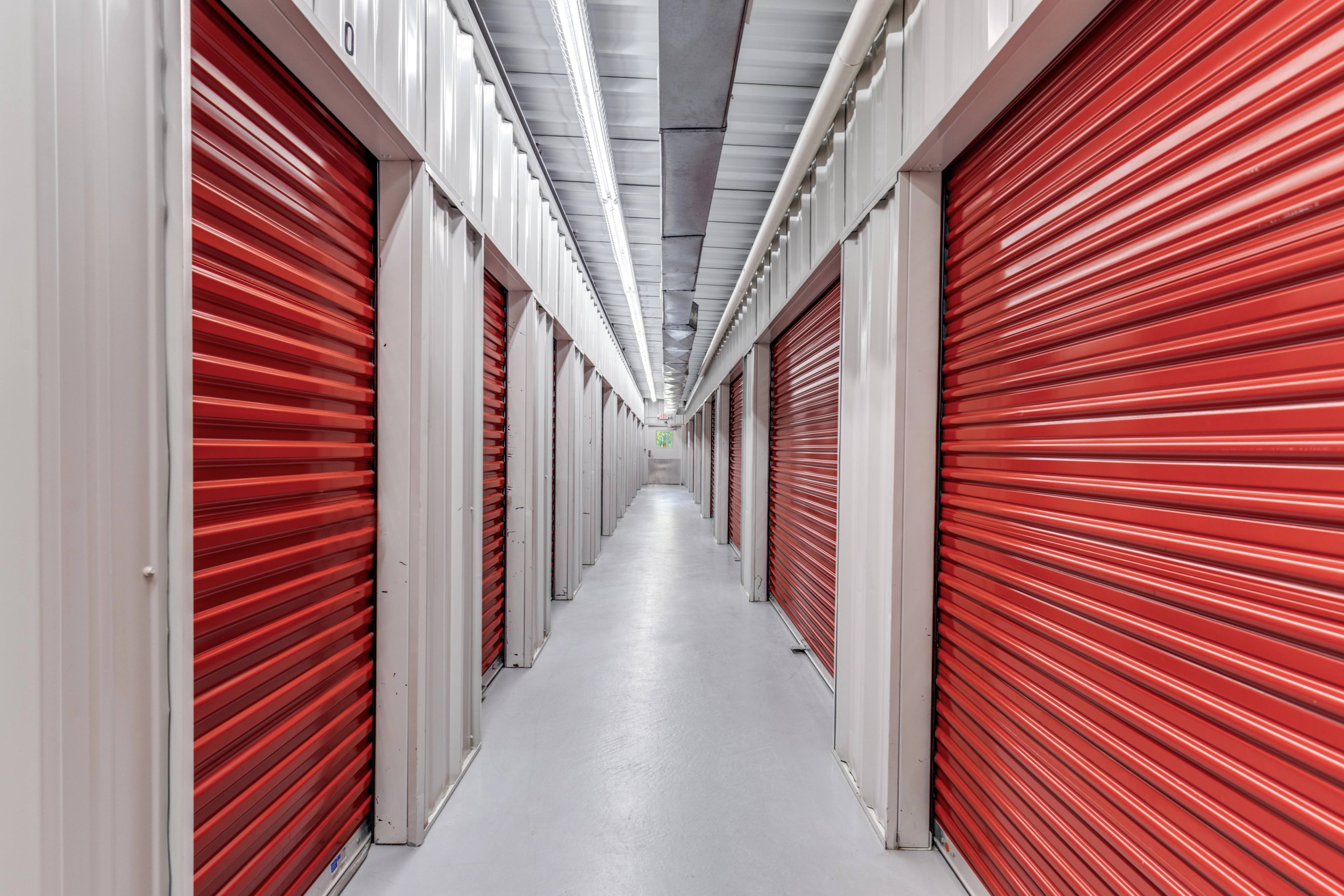 Interior corridor of a climate-controlled self-storage facility in Alvin, TX, lined with bright red metal roll-up doors, white unit walls, and overhead fluorescent lighting receding into the distance.