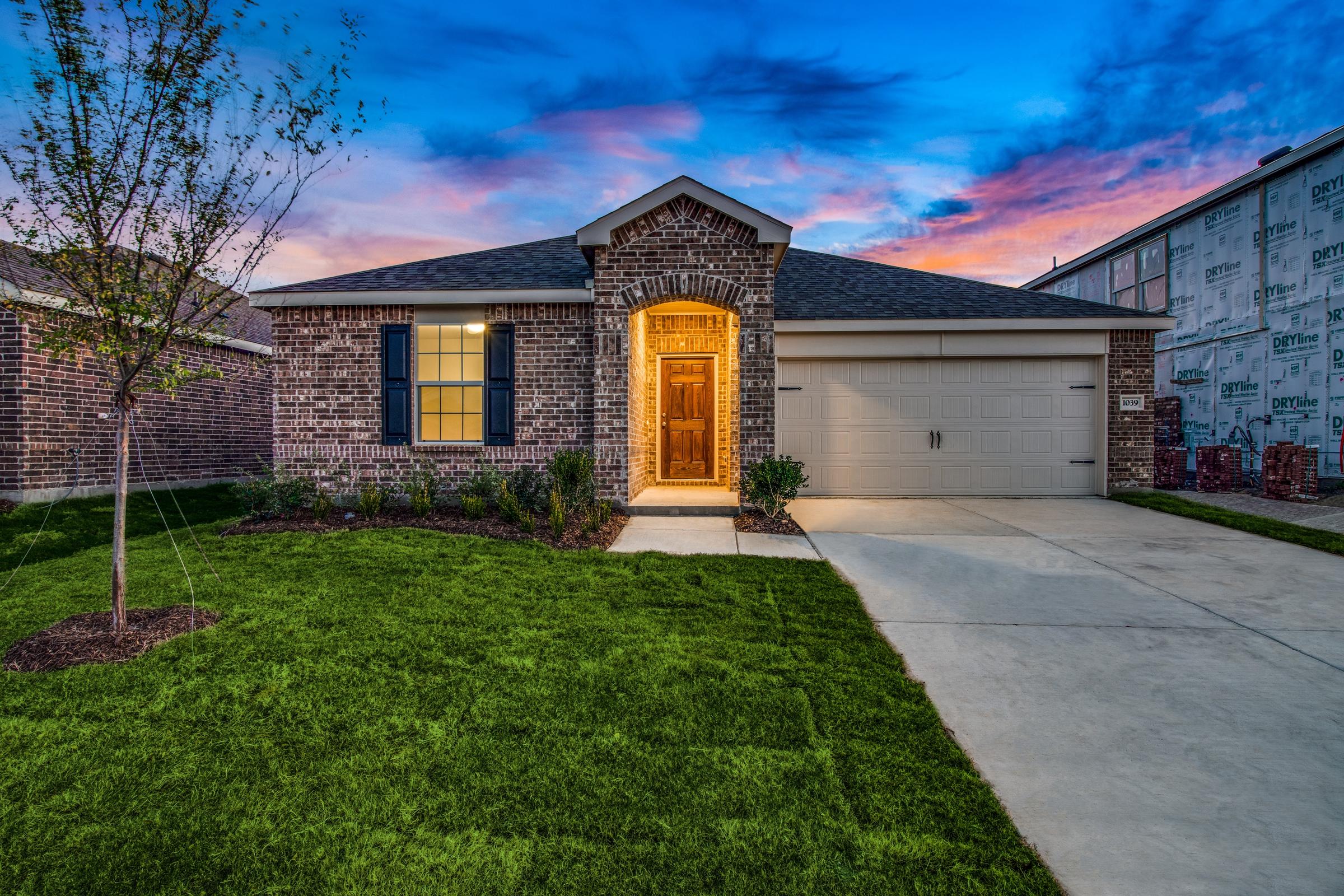 Brick spec home with warm porch light at twilight with a dramatic pink and blue sunset sky in a new construction community in Forney, Texas