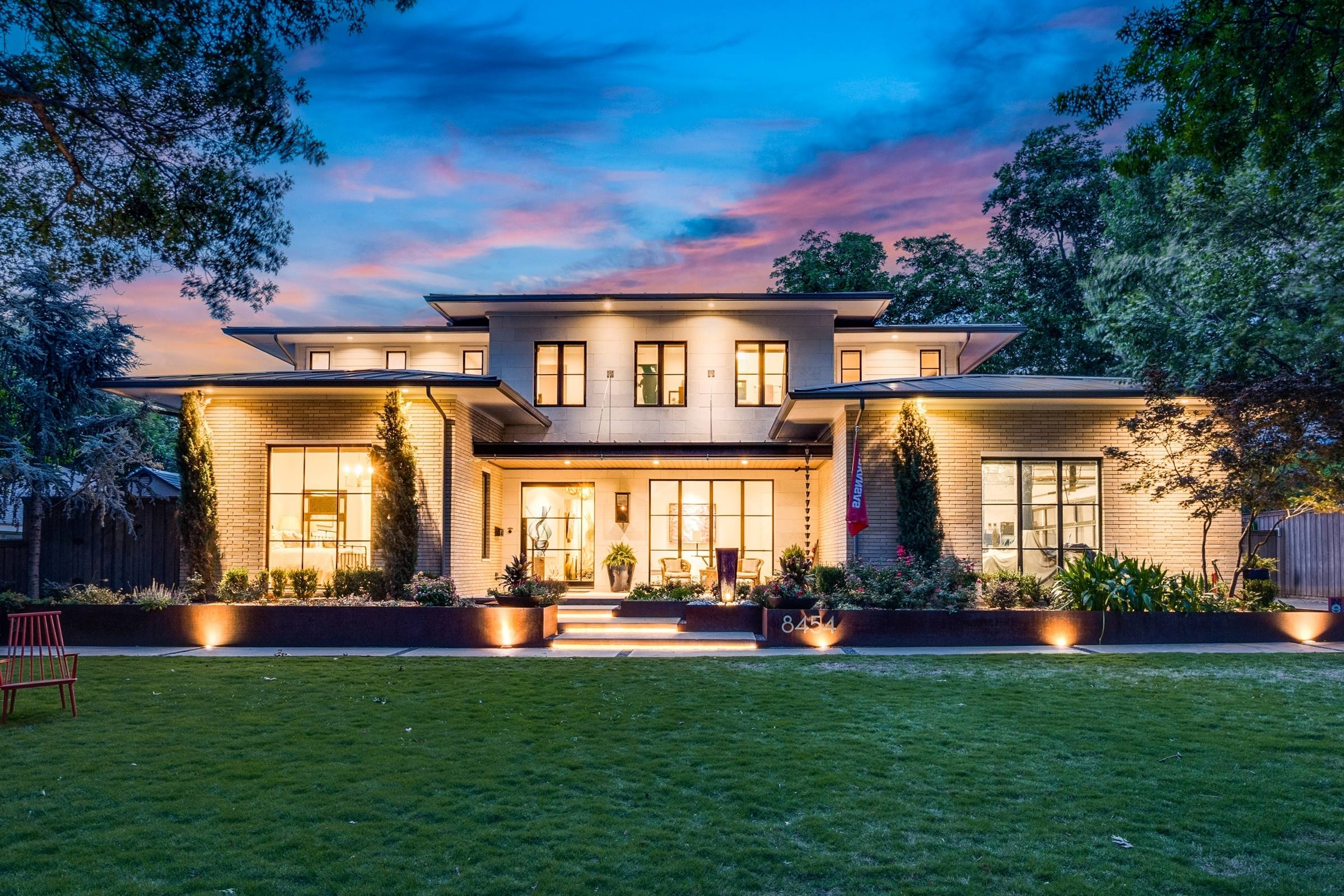 Modern prairie-style luxury home with warm interior lighting, manicured front lawn, and dramatic twilight sky in Dallas, Texas