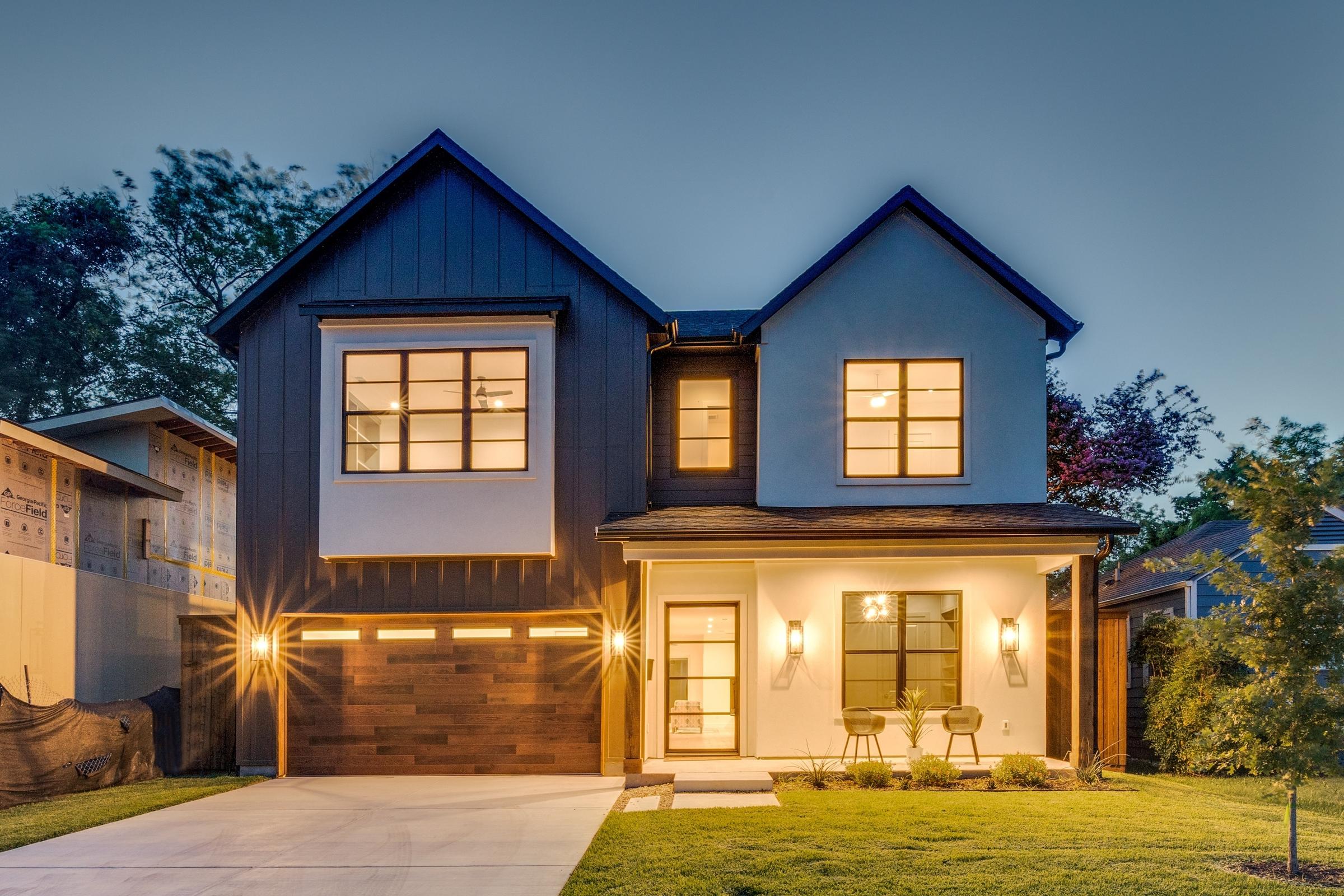 Two-story modern farmhouse-style new construction home with warm window glow and wood-accent garage door photographed at blue-hour twilight in Dallas, Texas
