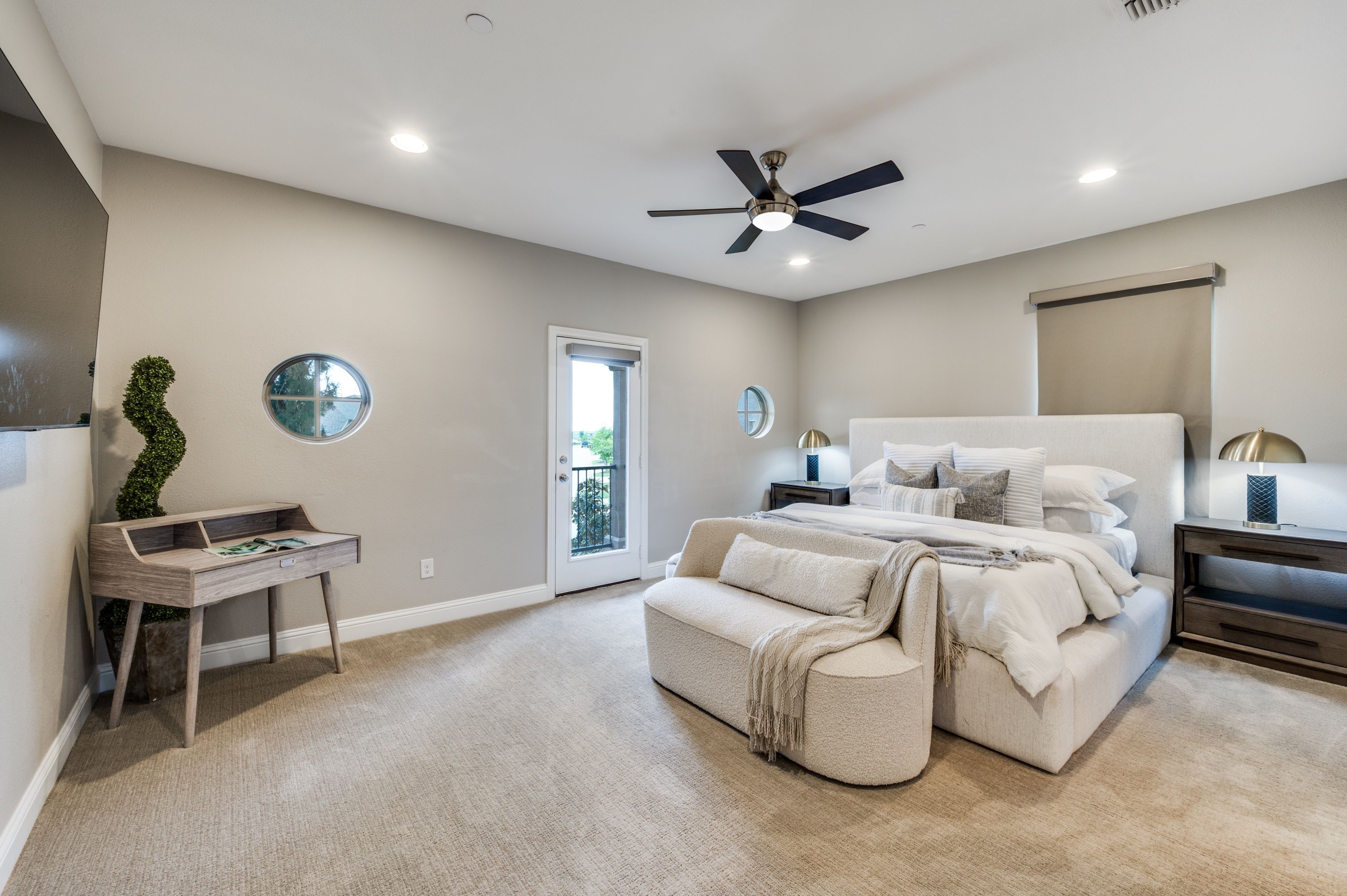 Light, neutral-toned master bedroom with ceiling fan on, upholstered bed, white sofa, and natural light from a balcony door, illustrating open blinds for photography prep.