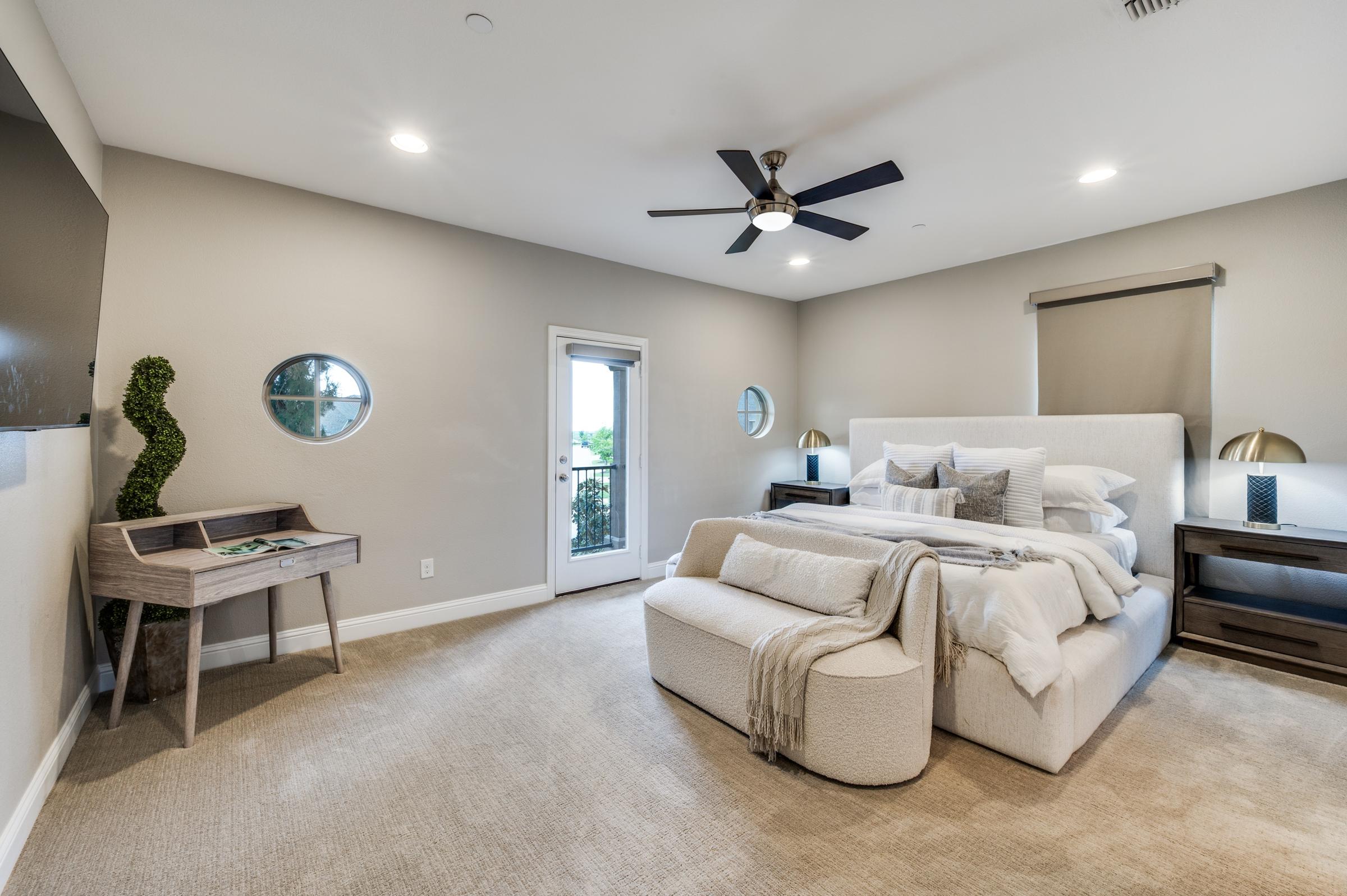 Light, neutral-toned master bedroom with ceiling fan on, upholstered bed, white sofa, and natural light from a balcony door, illustrating open blinds for photography prep.