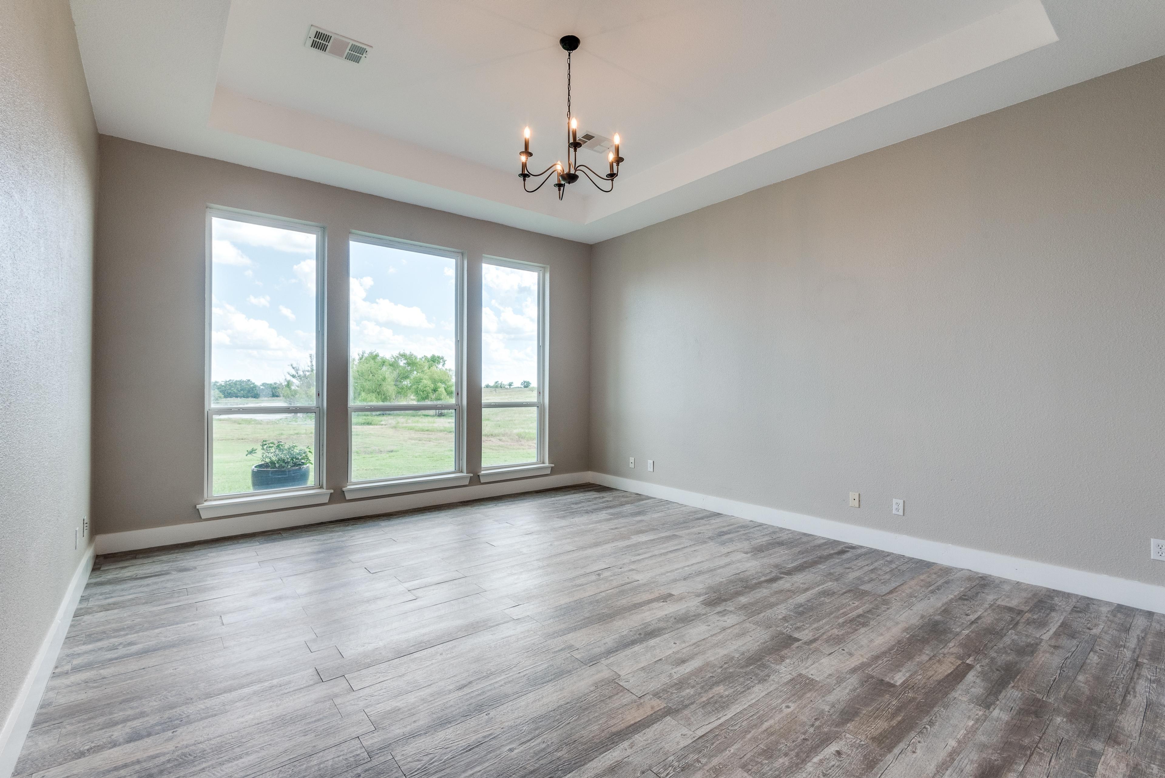 Empty master bedroom with tray ceiling, chandelier, and large windows overlooking countryside before virtual staging in Ennis, Texas