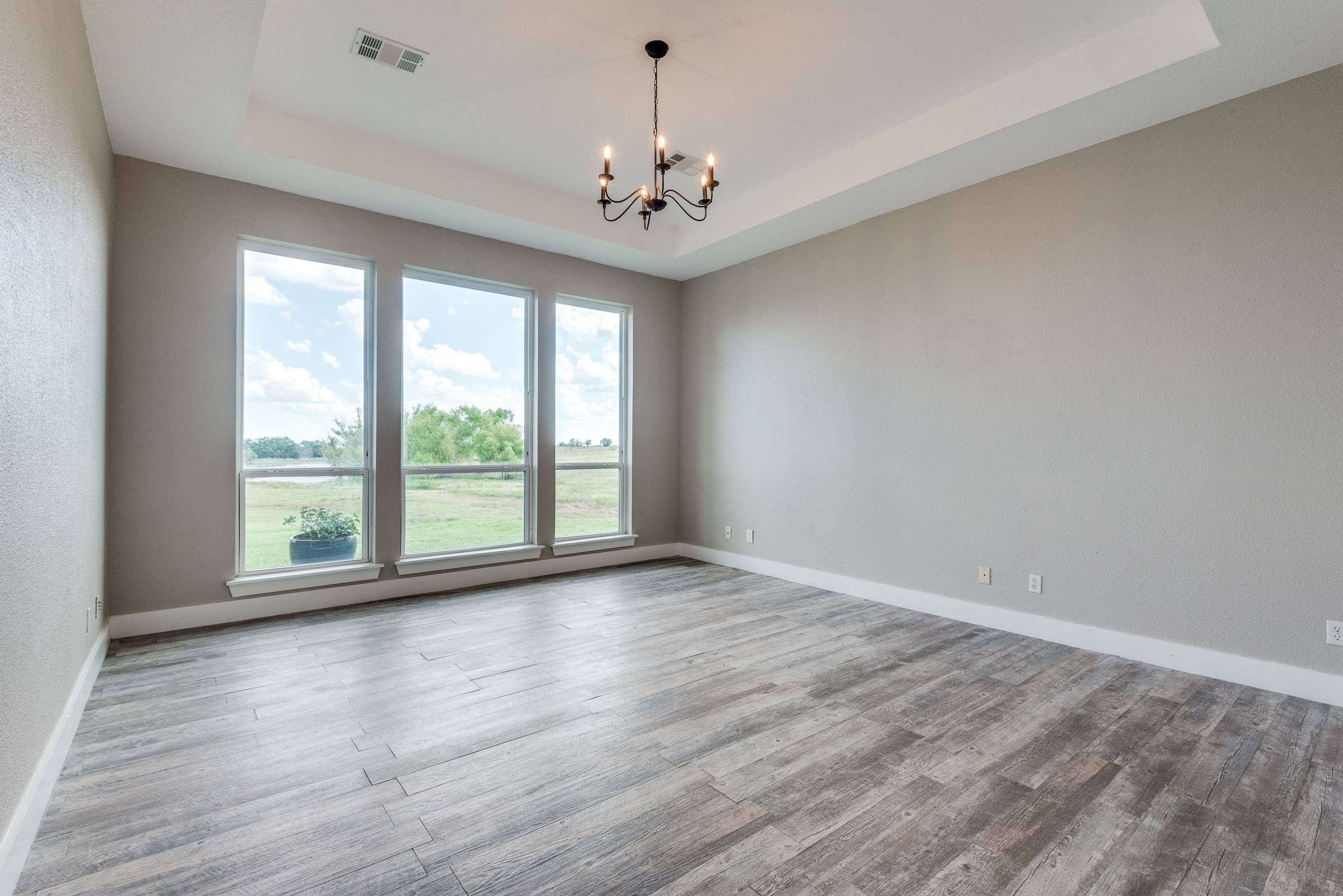 Empty master bedroom with tray ceiling, chandelier, and large windows overlooking countryside before virtual staging in Ennis, Texas