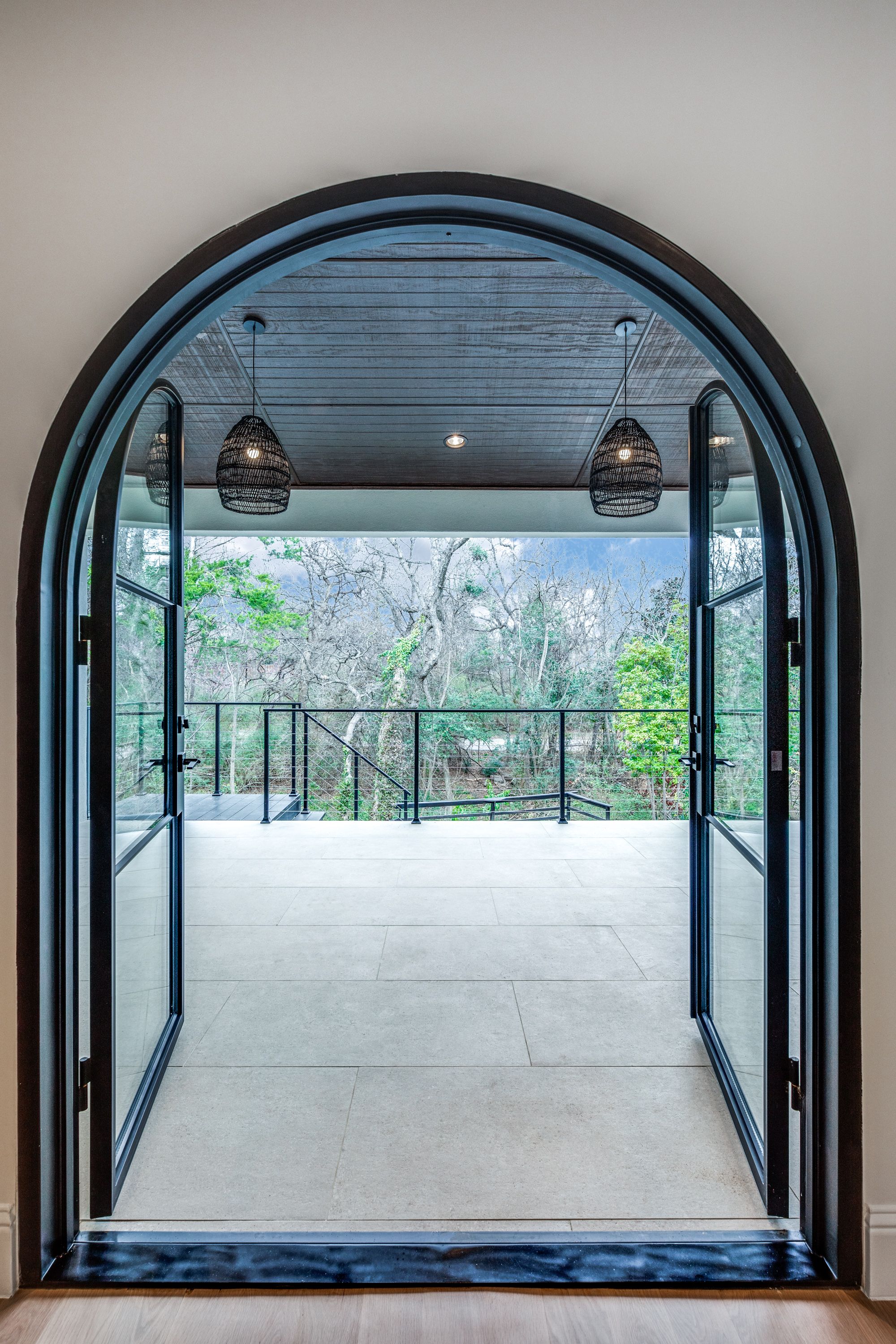Interior portrait view of an arched-top black steel-and-glass French door opening from a Keller, TX home to a covered patio with ceiling pendants and treetop views.