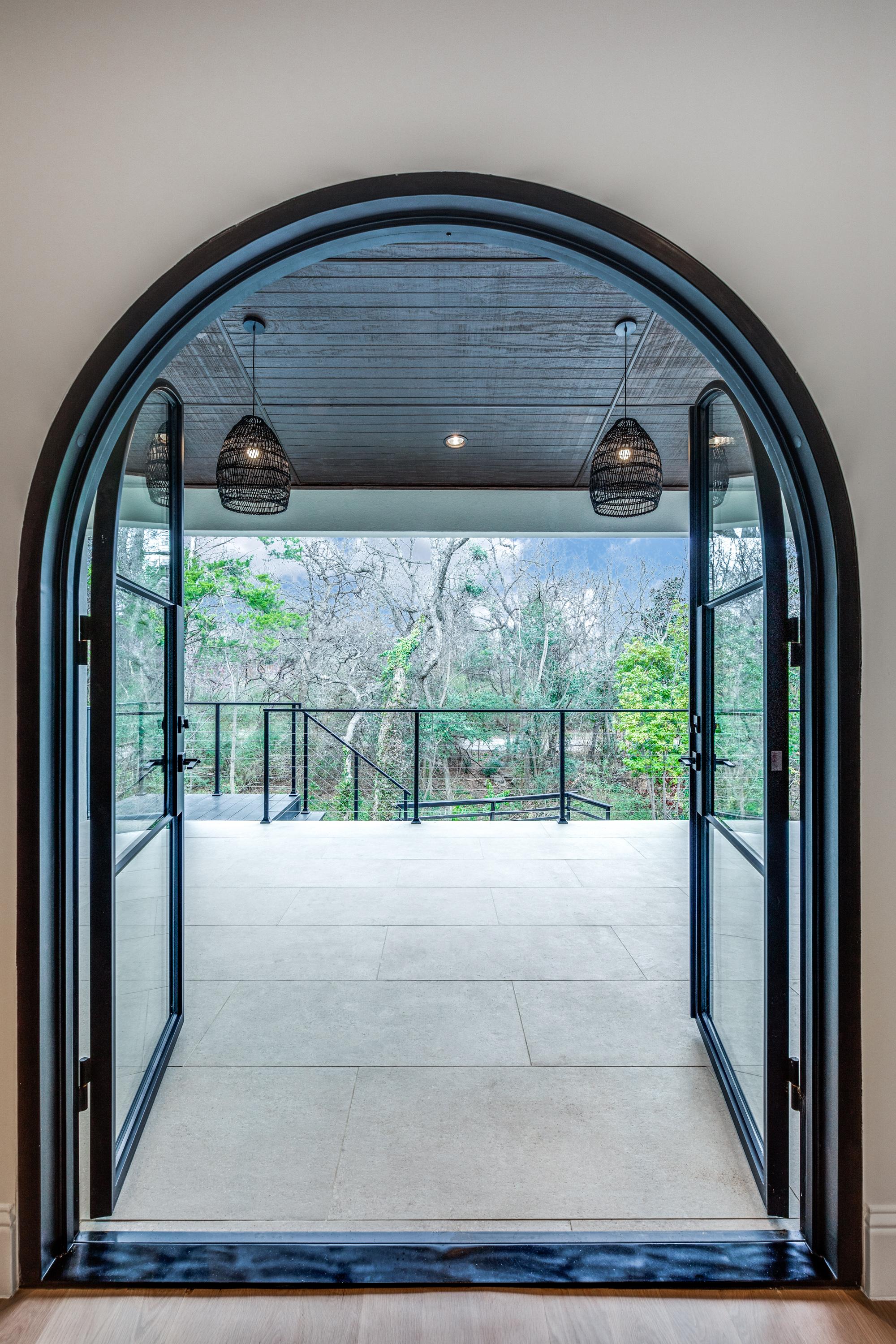 Interior portrait view of an arched-top black steel-and-glass French door opening from a Keller, TX home to a covered patio with ceiling pendants and treetop views.