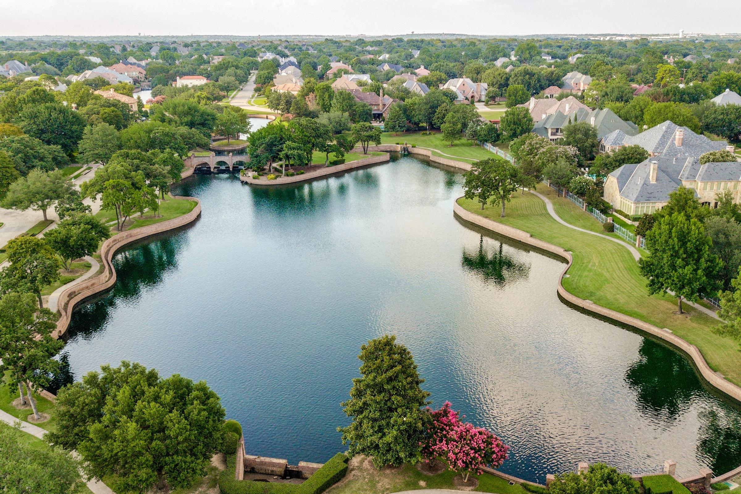 Aerial drone photograph of a luxury lakefront neighborhood with man-made lake, stone bridge, walking paths, and estate homes surrounded by mature trees in Colleyville, Texas