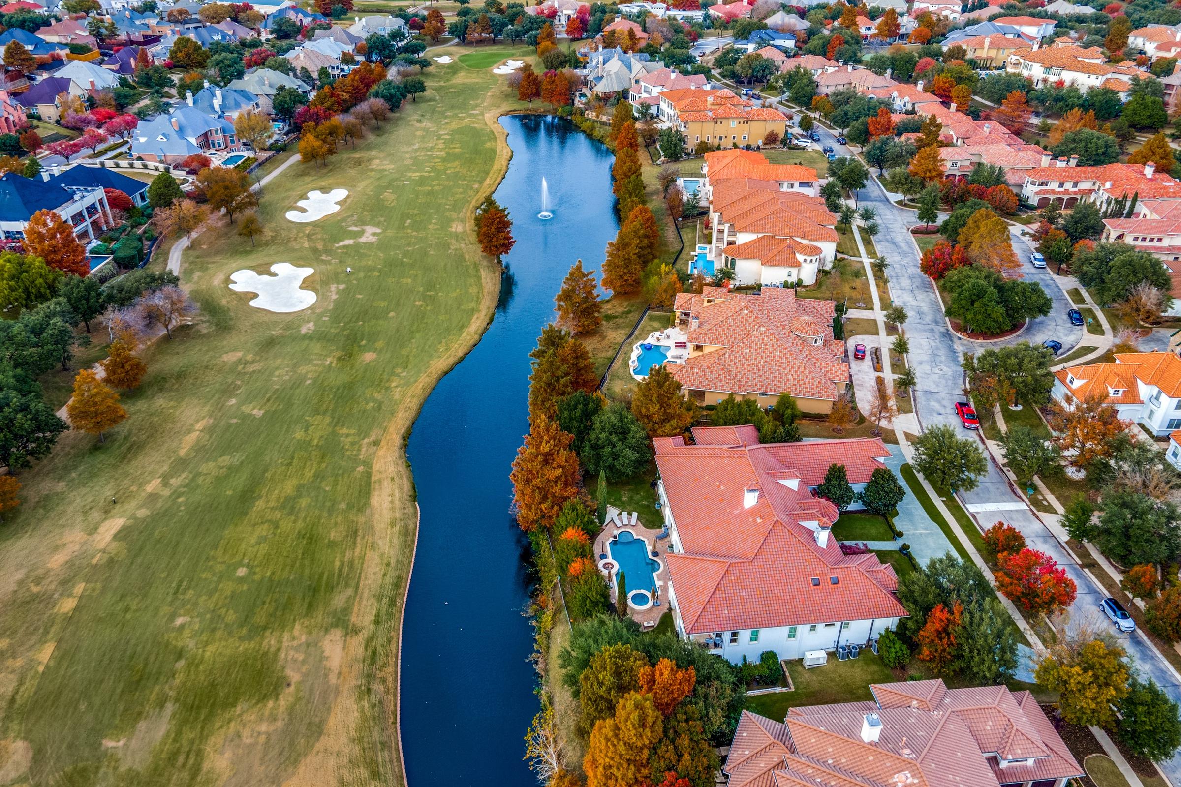 Aerial drone view of luxury Mediterranean-style homes along a golf course with autumn foliage and water feature in Irving, Texas