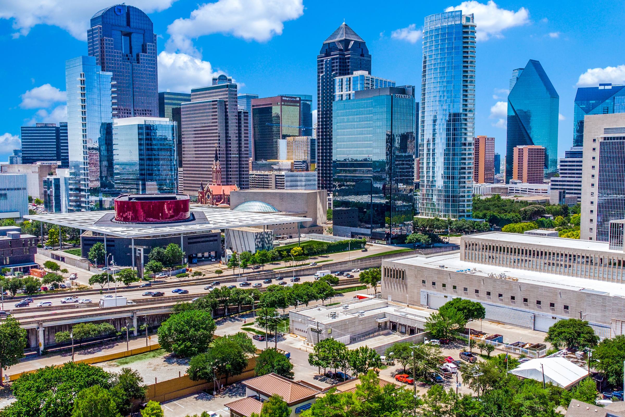 Aerial daytime view of the Dallas Arts District with the Winspear Opera House, downtown highrises, and Routh Street corridor under a bright blue sky, Dallas, Texas