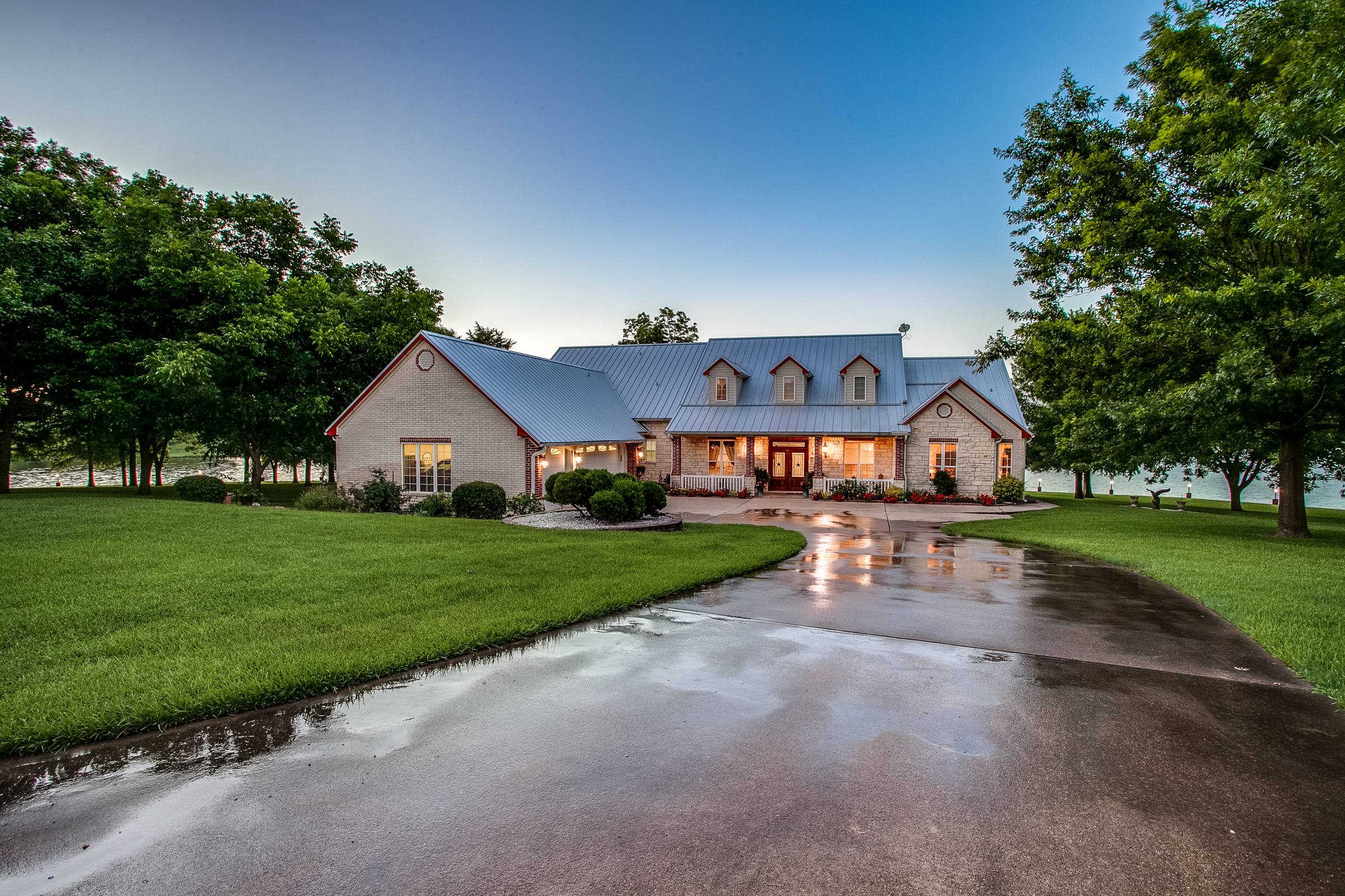 Texas country-style single-story home at twilight after rain, with warm interior lights glowing through windows, wraparound porch, and reflective wet driveway in Corsicana, TX