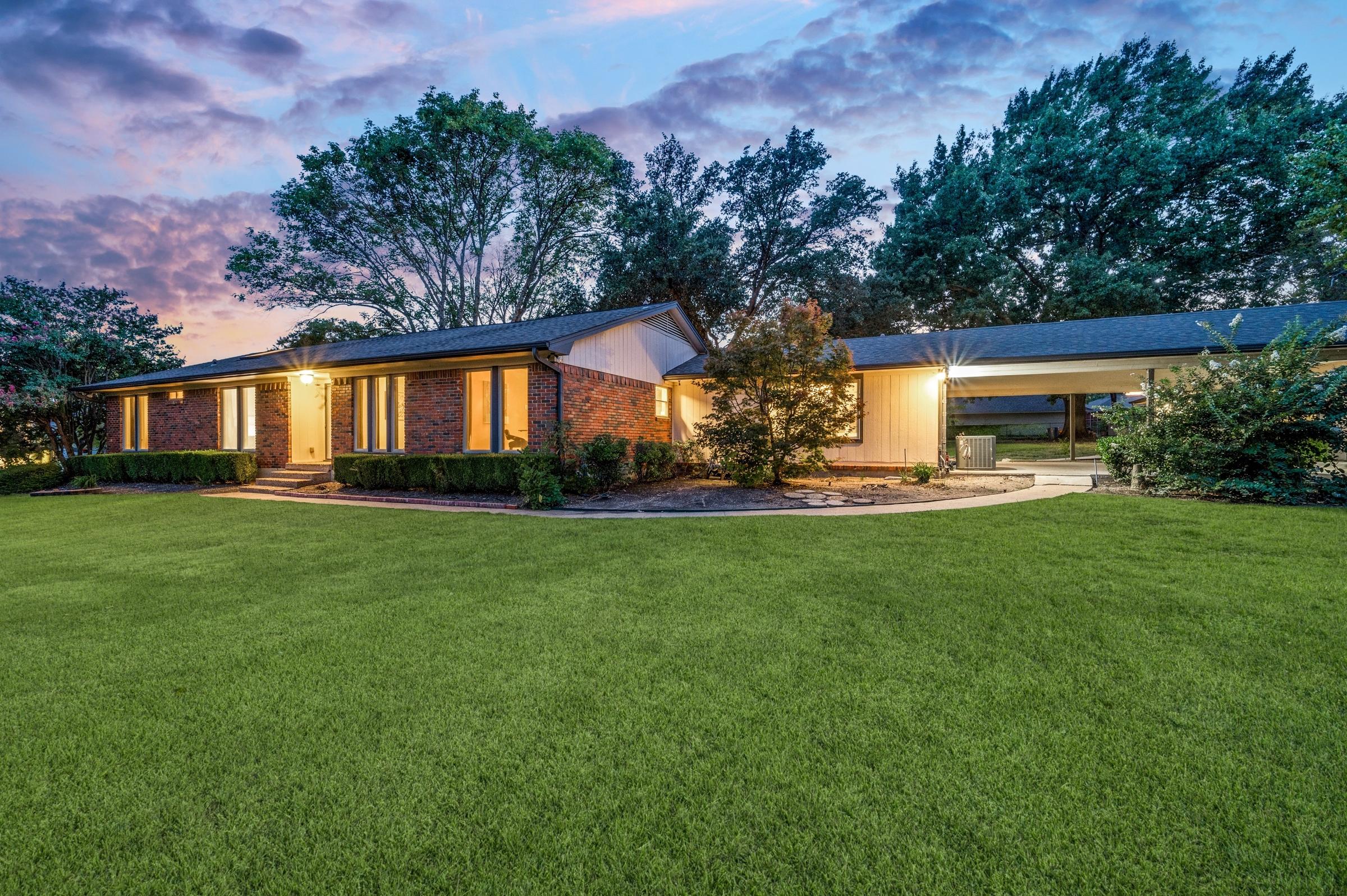 Single-story mid-century brick ranch home with warm window glow, manicured lawn, and mature trees at twilight in Dallas, Texas