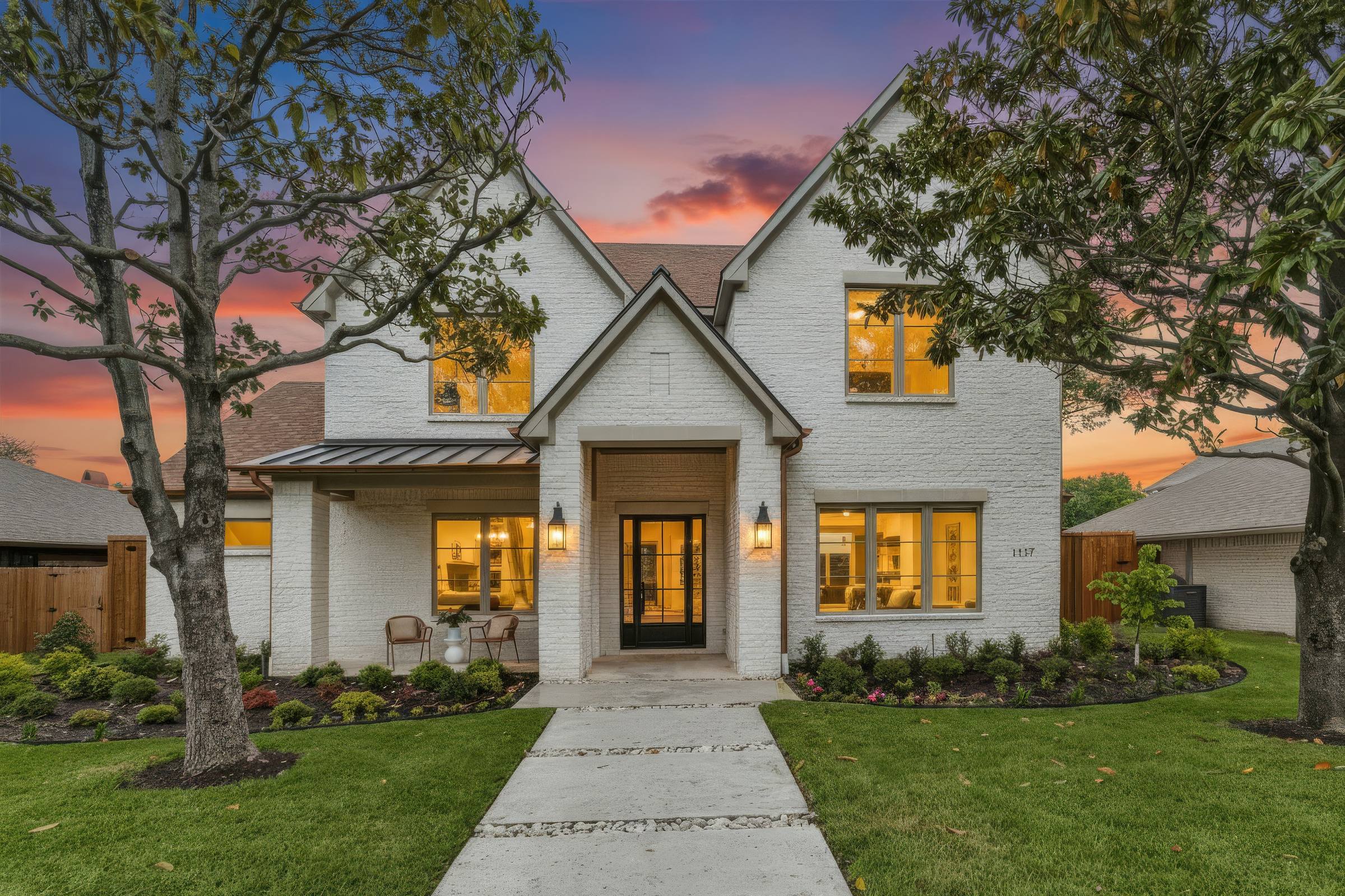 Digitally enhanced twilight exterior of a two-story white-brick transitional home in Richardson, TX, with warm window glow, landscape lighting, manicured beds, and a saturated purple-and-pink dusk sky.