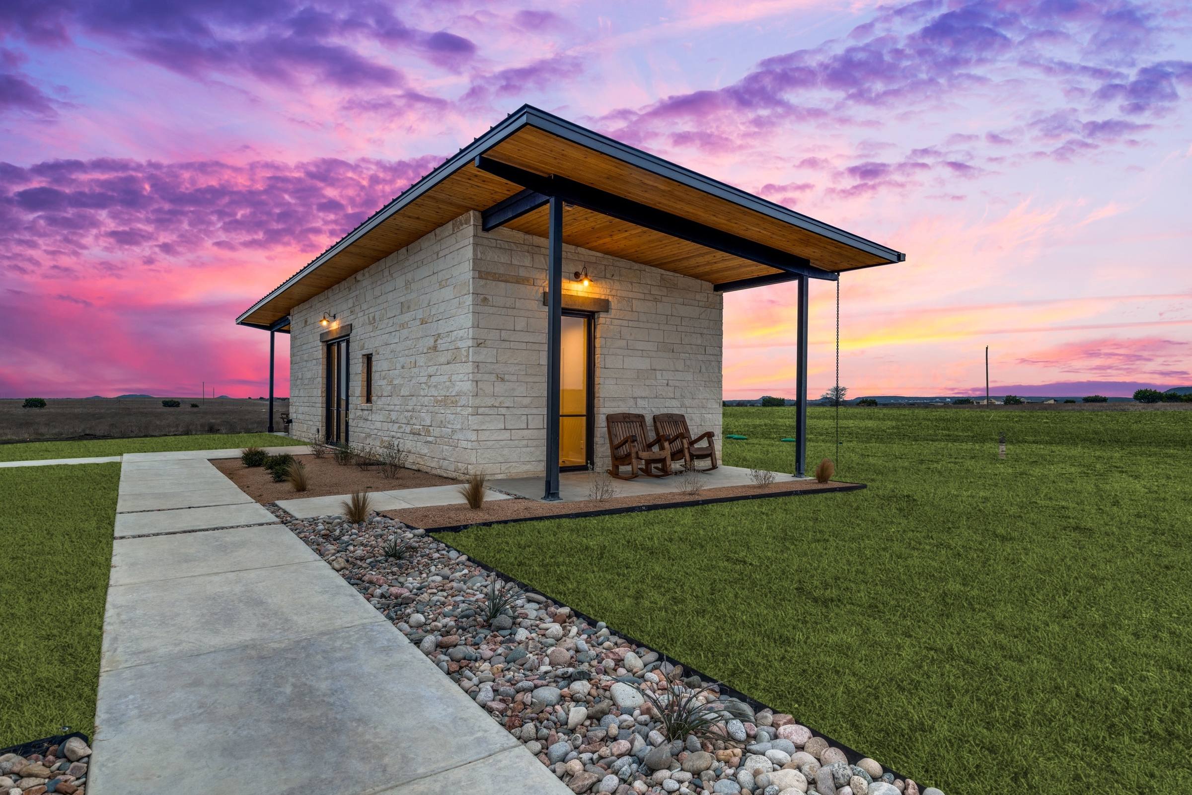 Modern limestone ADU guest house with covered porch and rocking chairs against a dramatic pink and purple twilight sky in Iredell, Texas