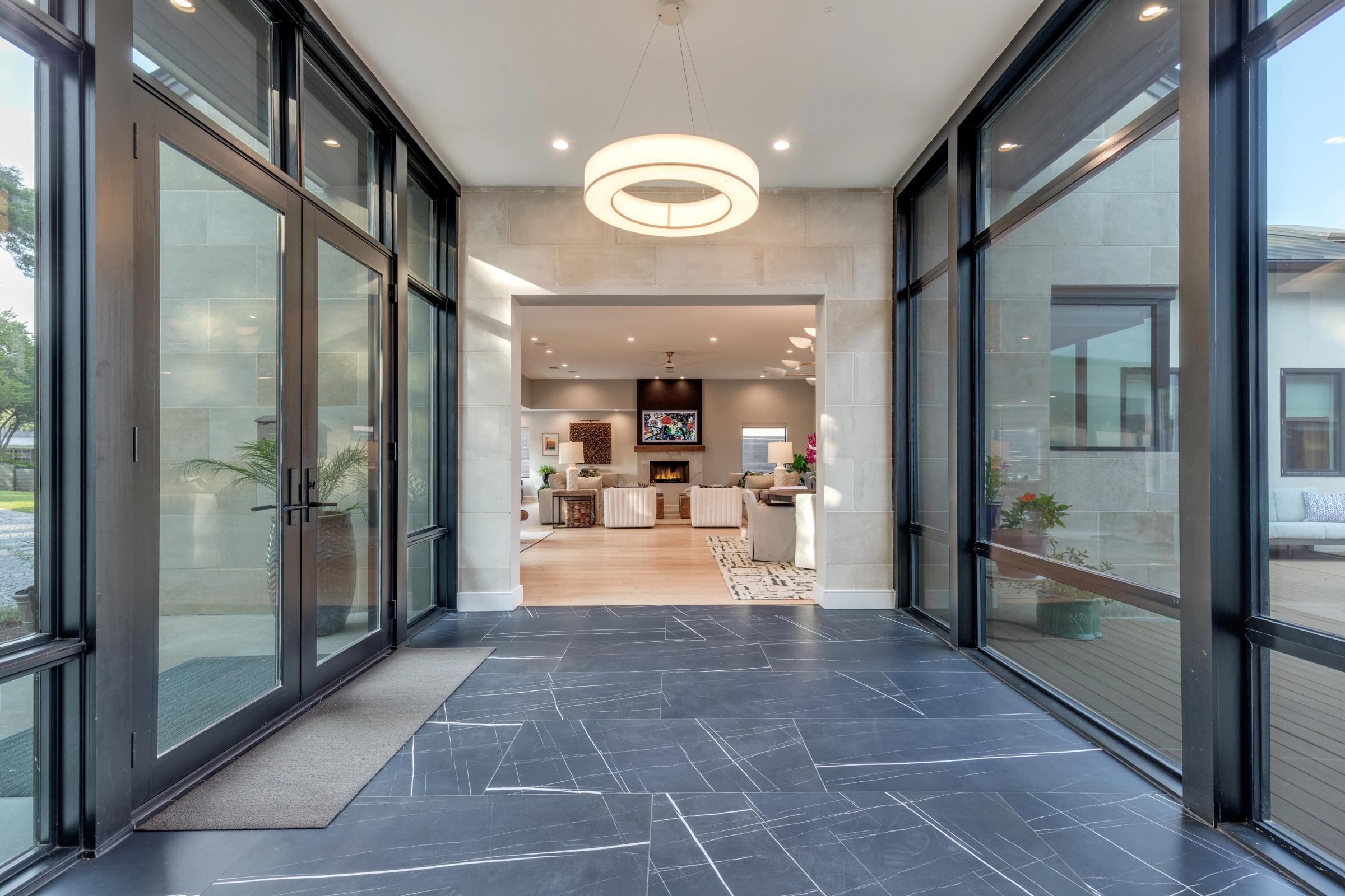 Modern glass-walled entryway with geometric slate floor, circular pendant light, and a view into the living room with fireplace in a luxury Dallas home.