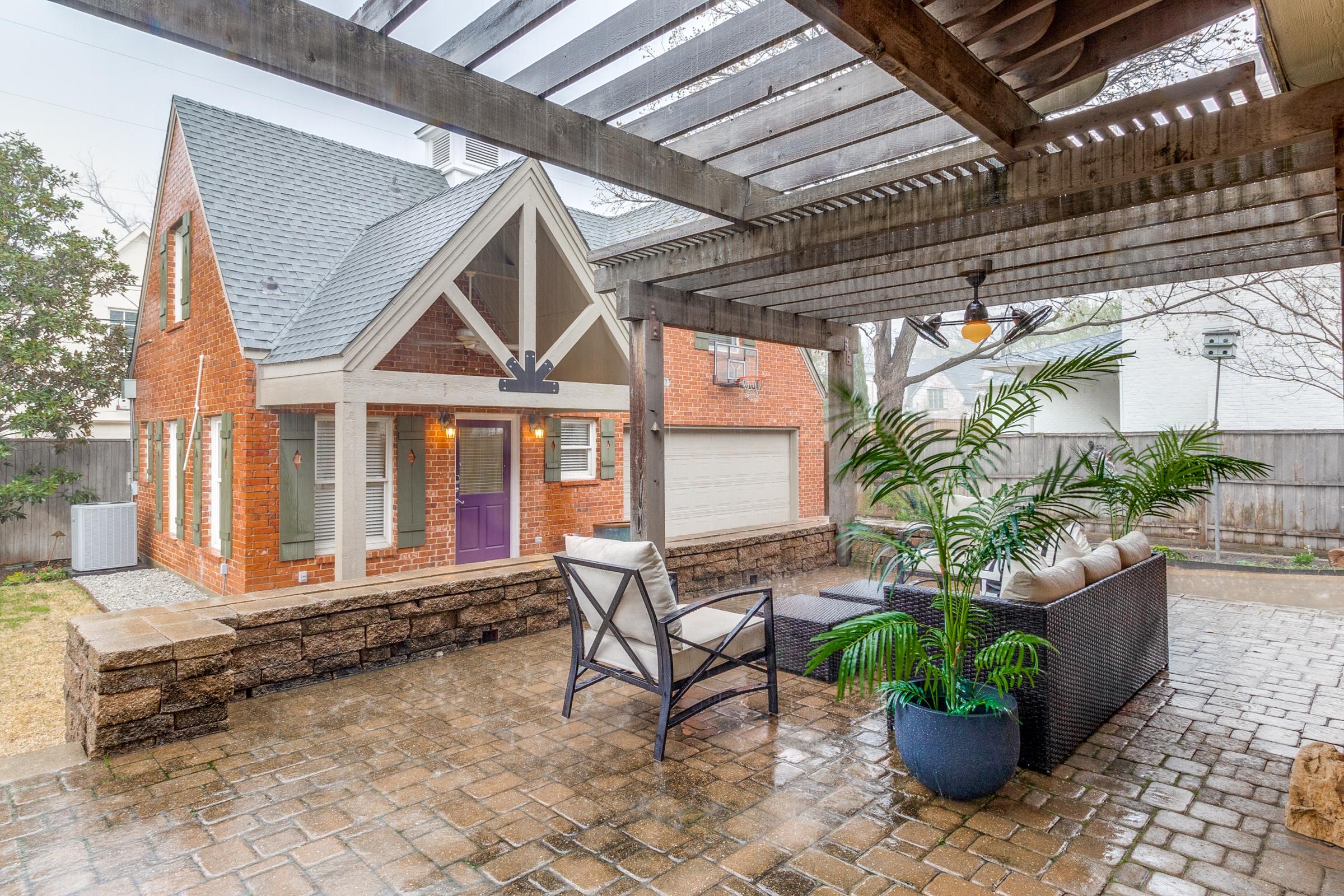 Covered patio with pergola, stone planter, and seating area outside a red-brick Dallas, TX home during heavy rain — showing that covered outdoor spaces can still be photographed when weather turns.