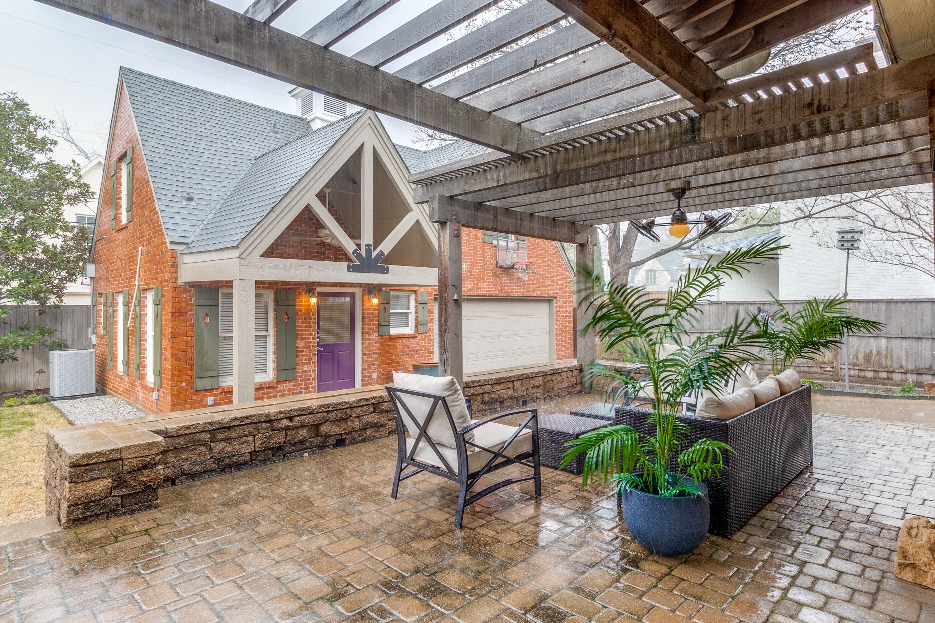 Covered patio with pergola, stone planter, and seating area outside a red-brick Dallas, TX home during heavy rain โ showing that covered outdoor spaces can still be photographed when weather turns.