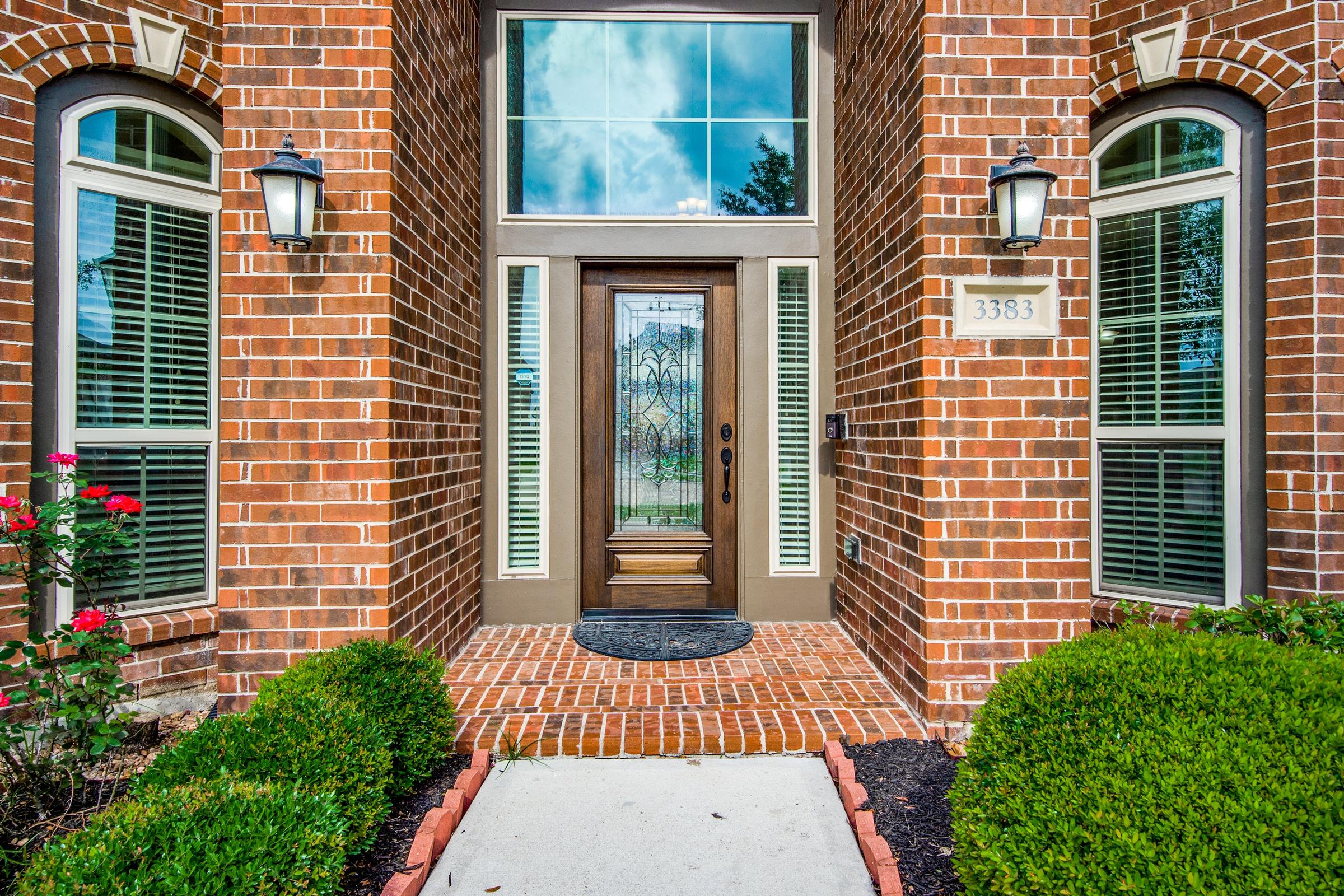 Traditional red brick home entrance with stained glass front door, arched windows, lantern sconces, and manicured boxwood hedges in Pearland, Texas