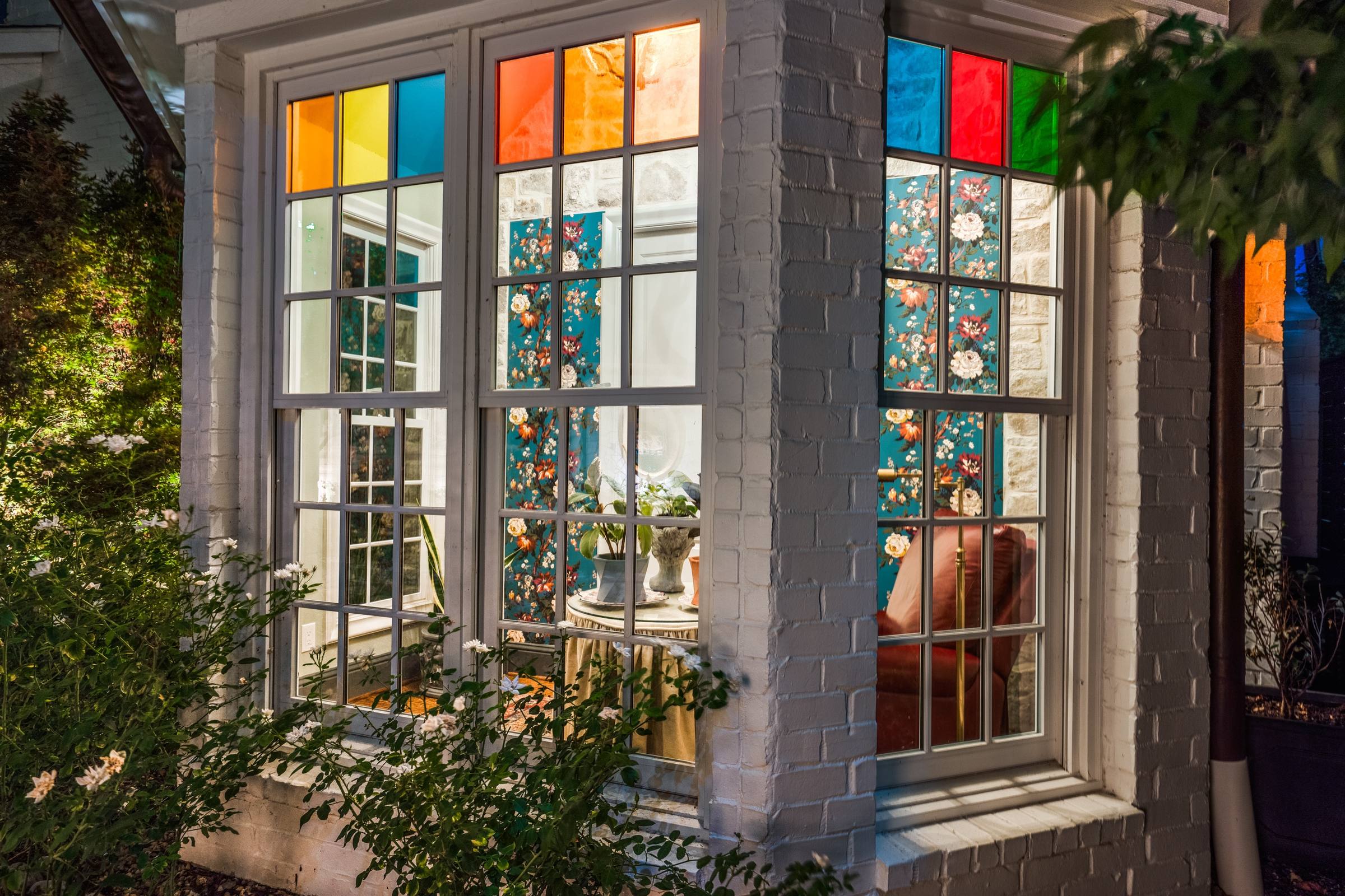 Evening twilight view of a painted brick bay window with colorful stained glass panels framing a floral wallpapered breakfast nook at a historic Lakehurst estate.